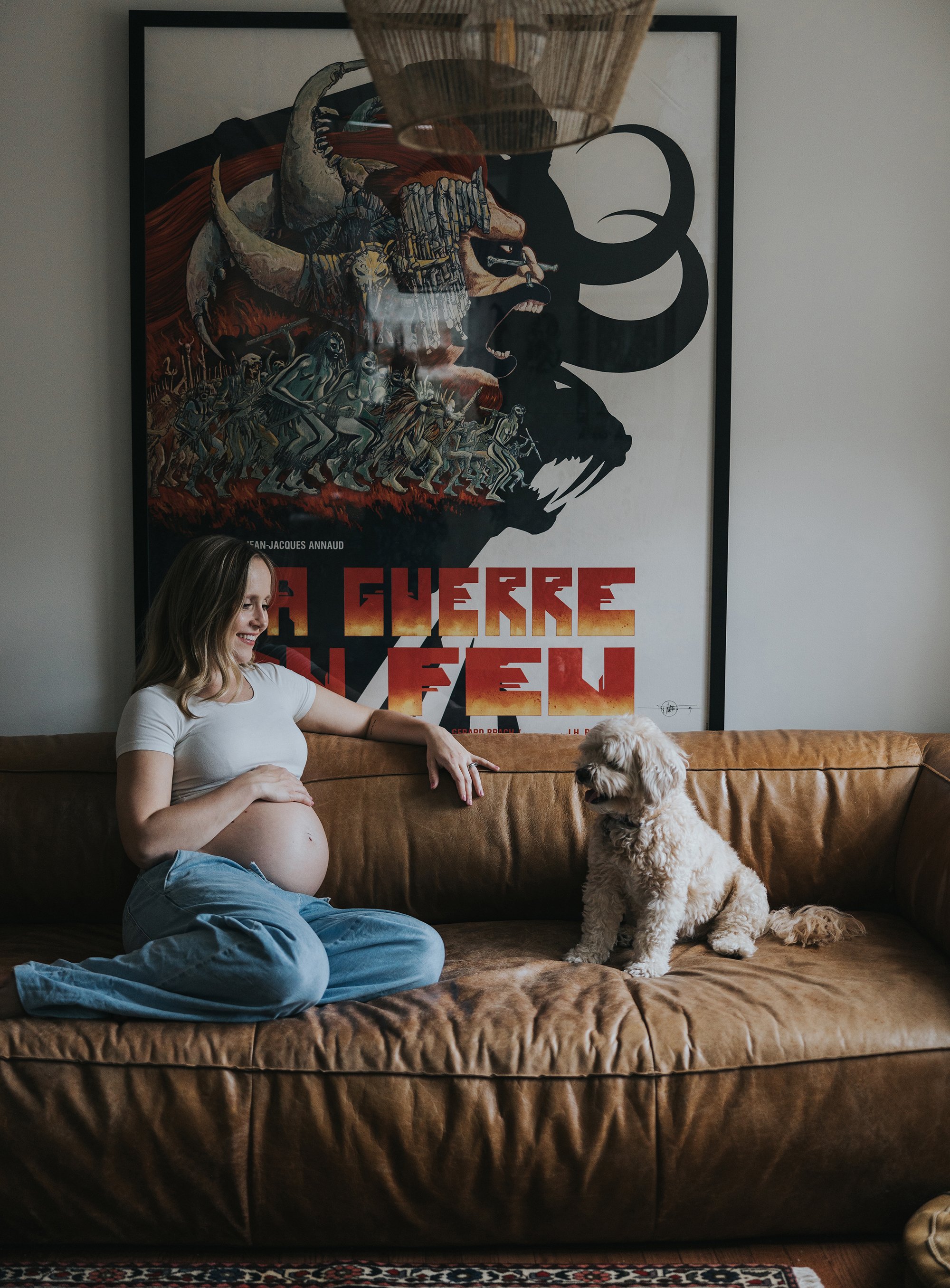 A pregnant woman sitting on a leather sofa, looking at her small dog, with a large artistic poster on the wall behind her, in a stylish home interior.