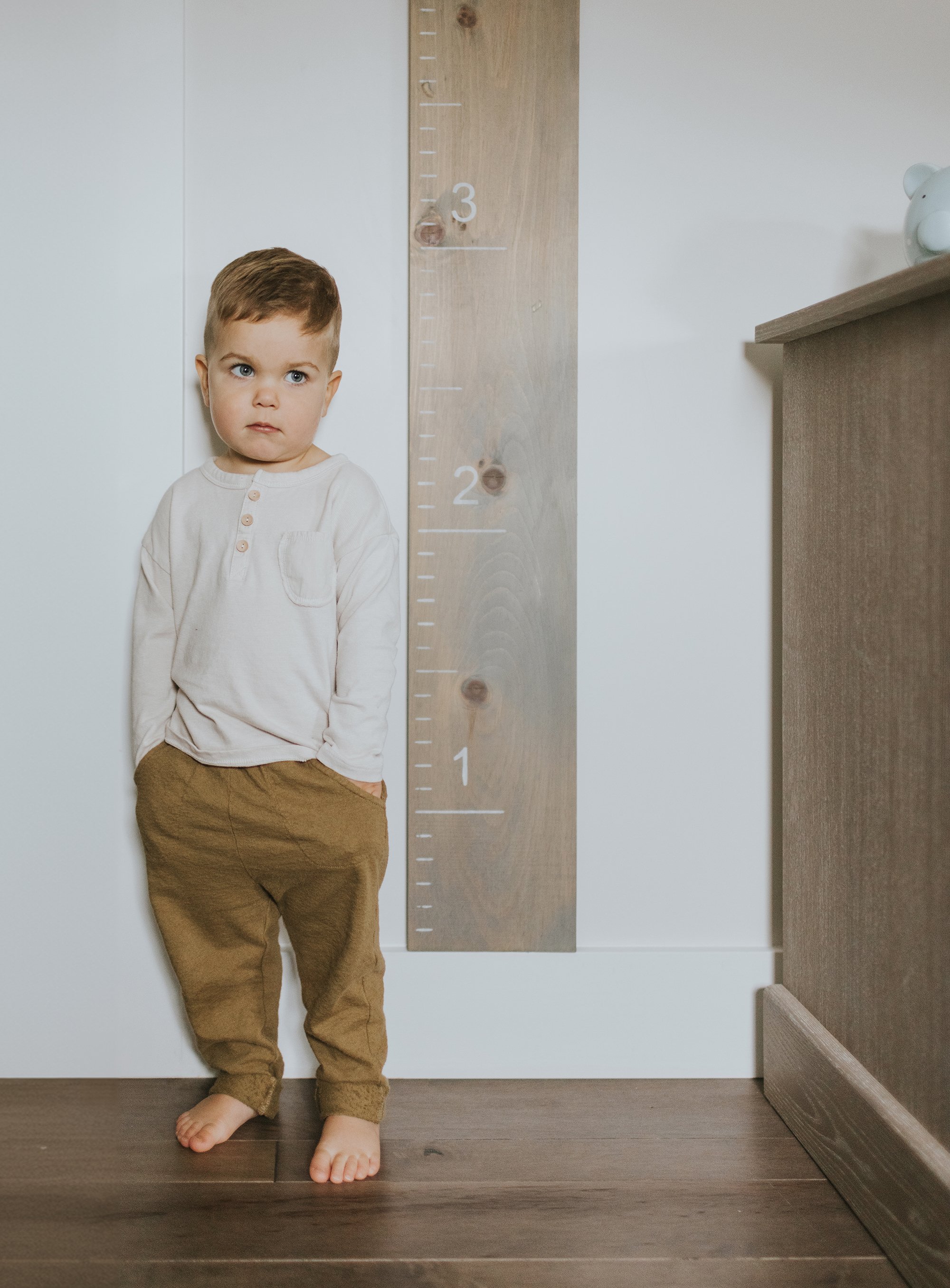 A toddler stands against a growth chart on the wall, looking thoughtful with hands in pockets.