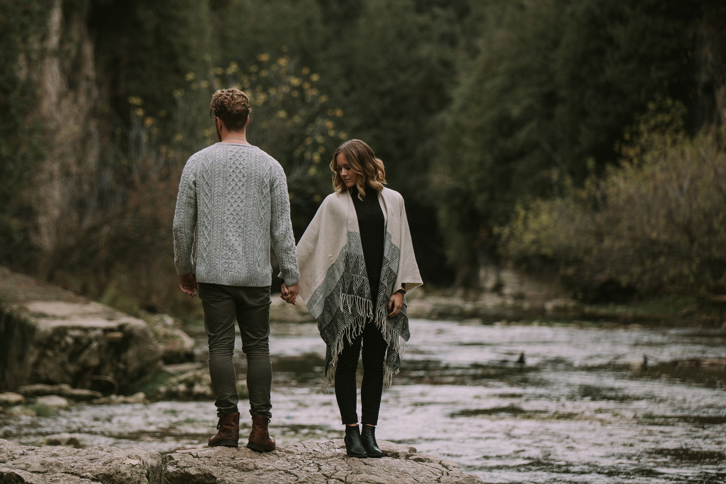 A couple holding hands while standing on rocks by a river, dressed in cozy sweaters, capturing the essence of a rustic fall engagement session.