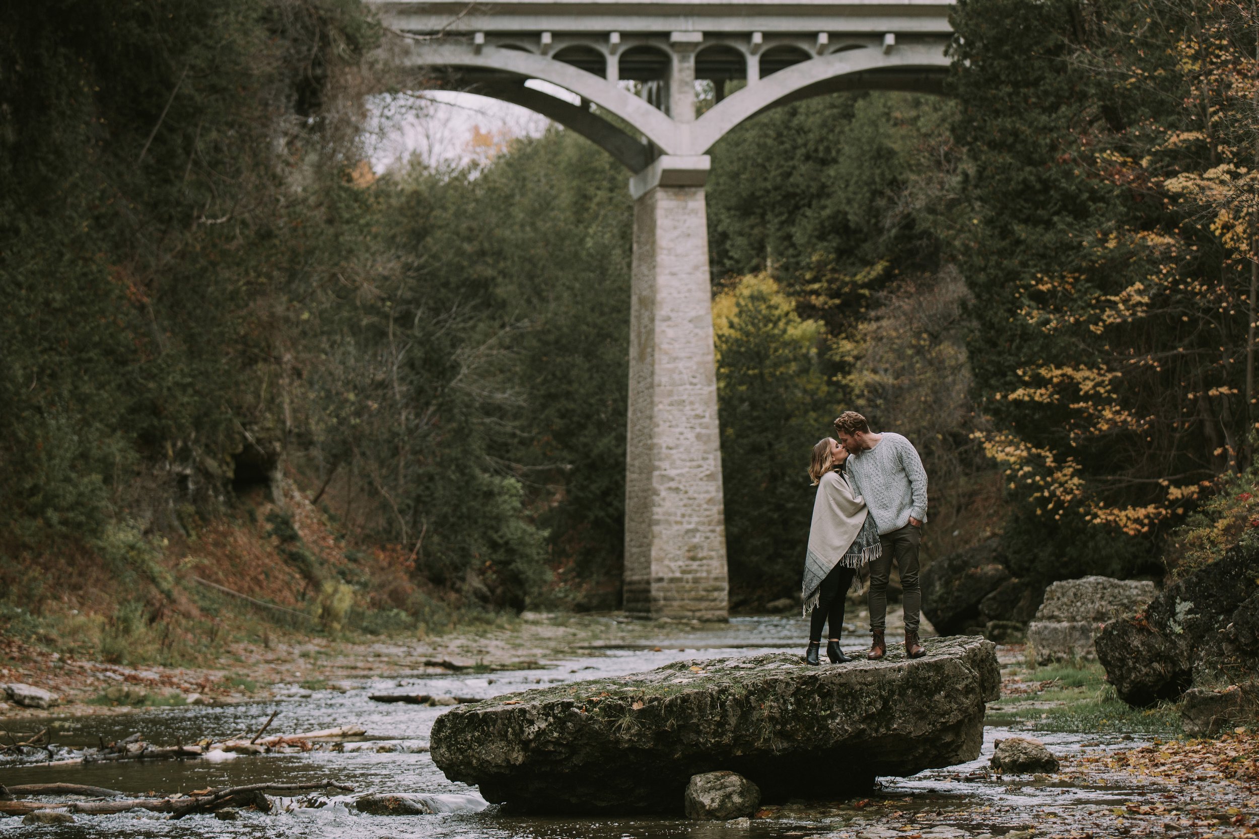 A couple holds hands while standing on a large rock beneath a bridge during an autumn engagement session, surrounded by a rustic, natural landscape.