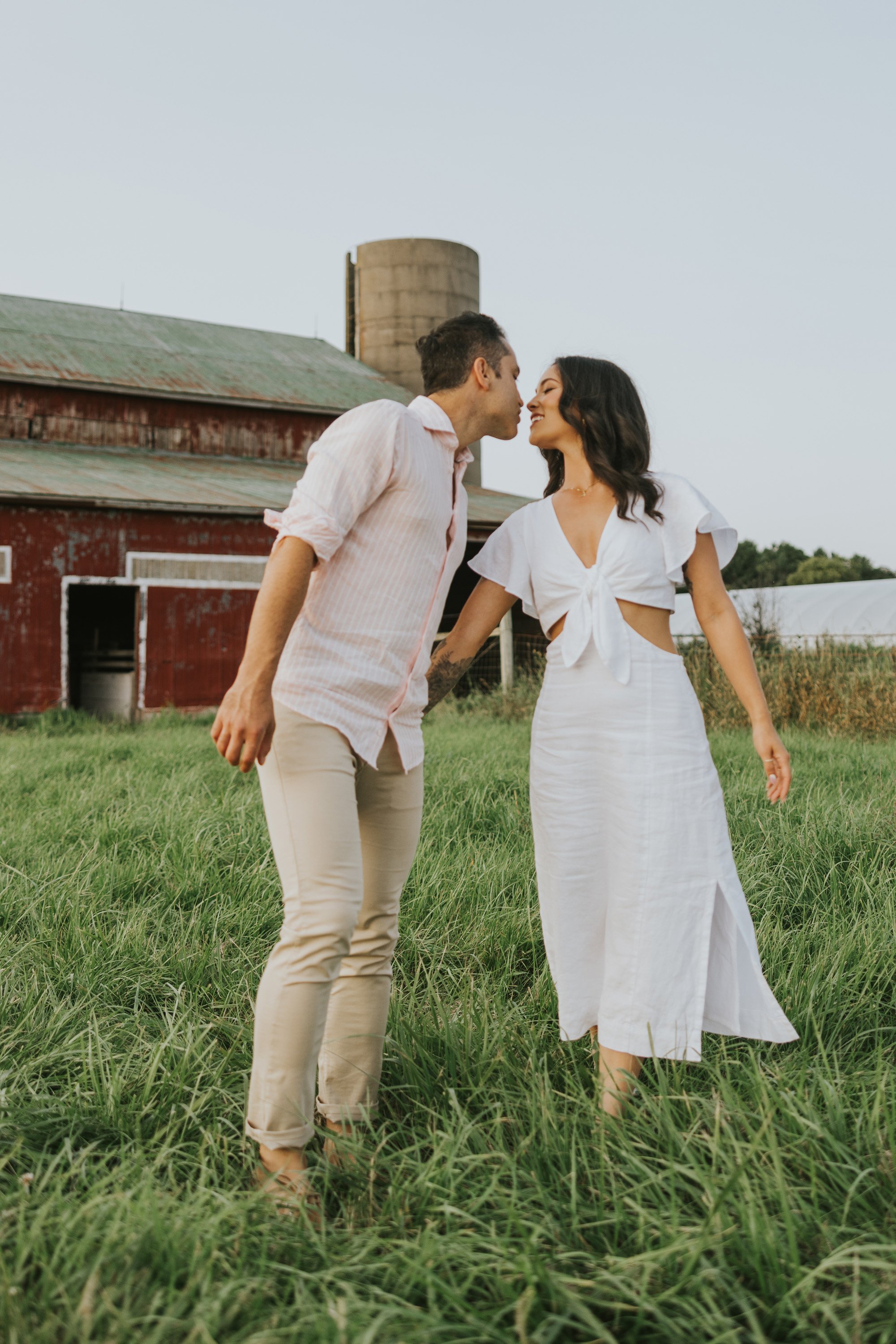Couple sharing a kiss near a farm silo, surrounded by green pasture, during their engagement session at sunset.