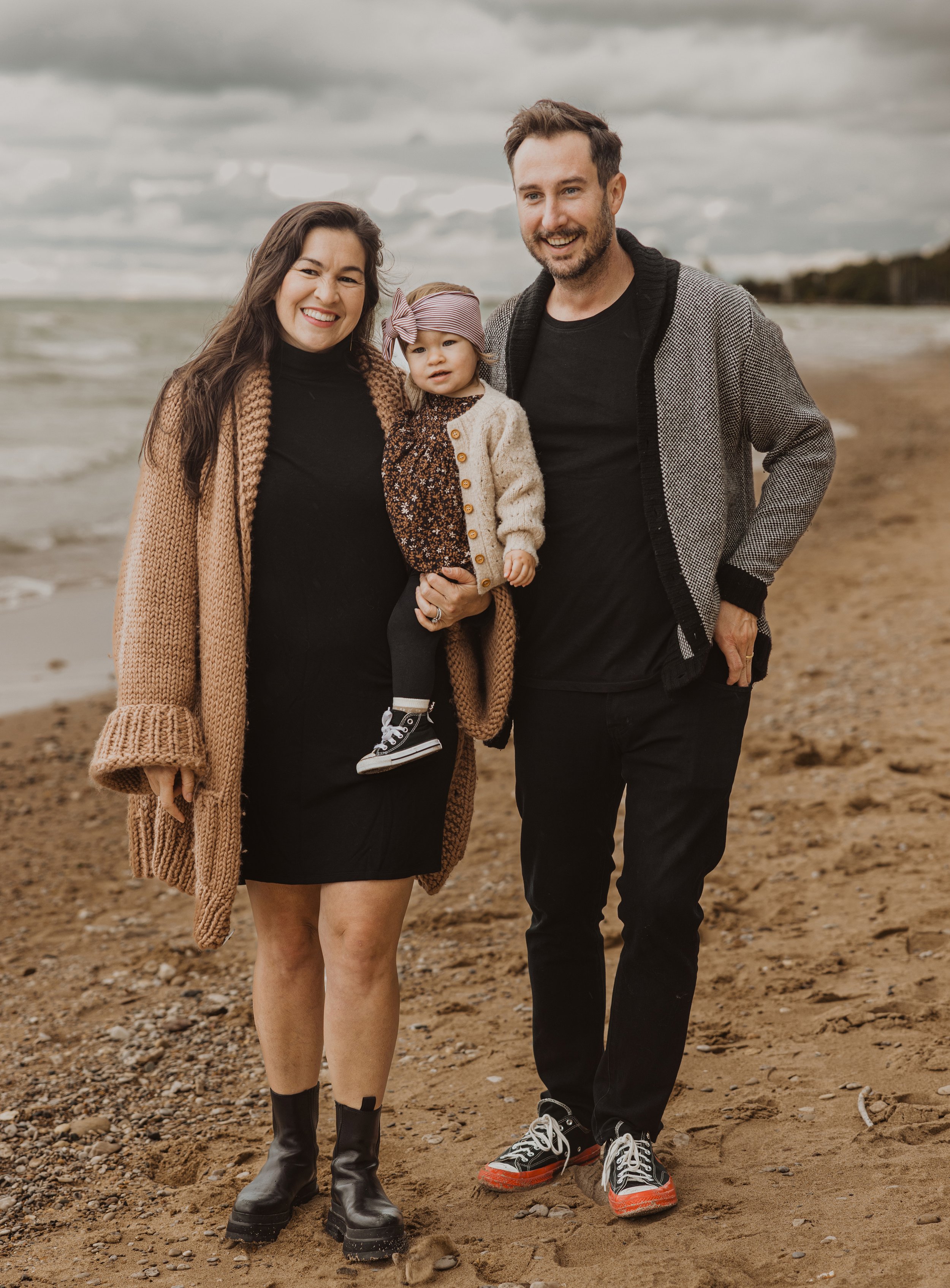 A family portrait on a moody beach with parents holding their young daughter dressed warmly against the cloudy weather.