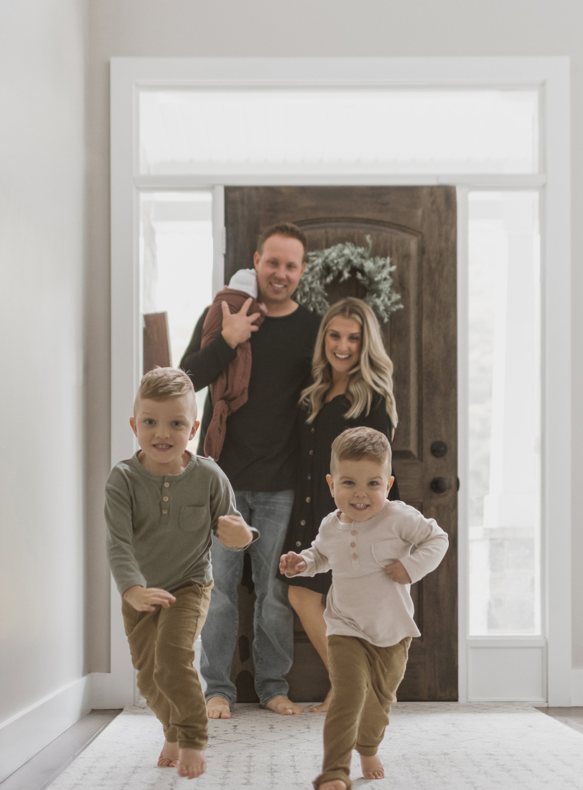 A joyful family welcoming home their new baby, with two young boys running towards the camera and parents smiling in the background.