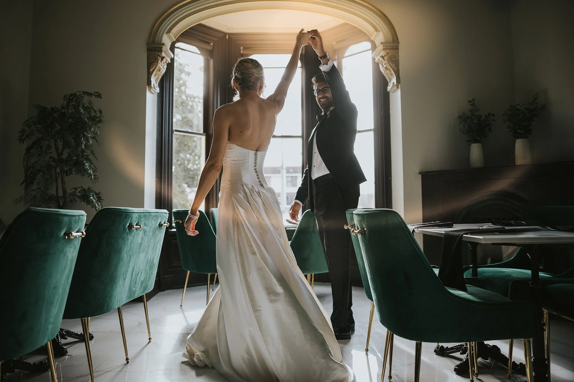 A bride and groom share a joyful dance in an elegantly decorated room with large windows and green velvet chairs.