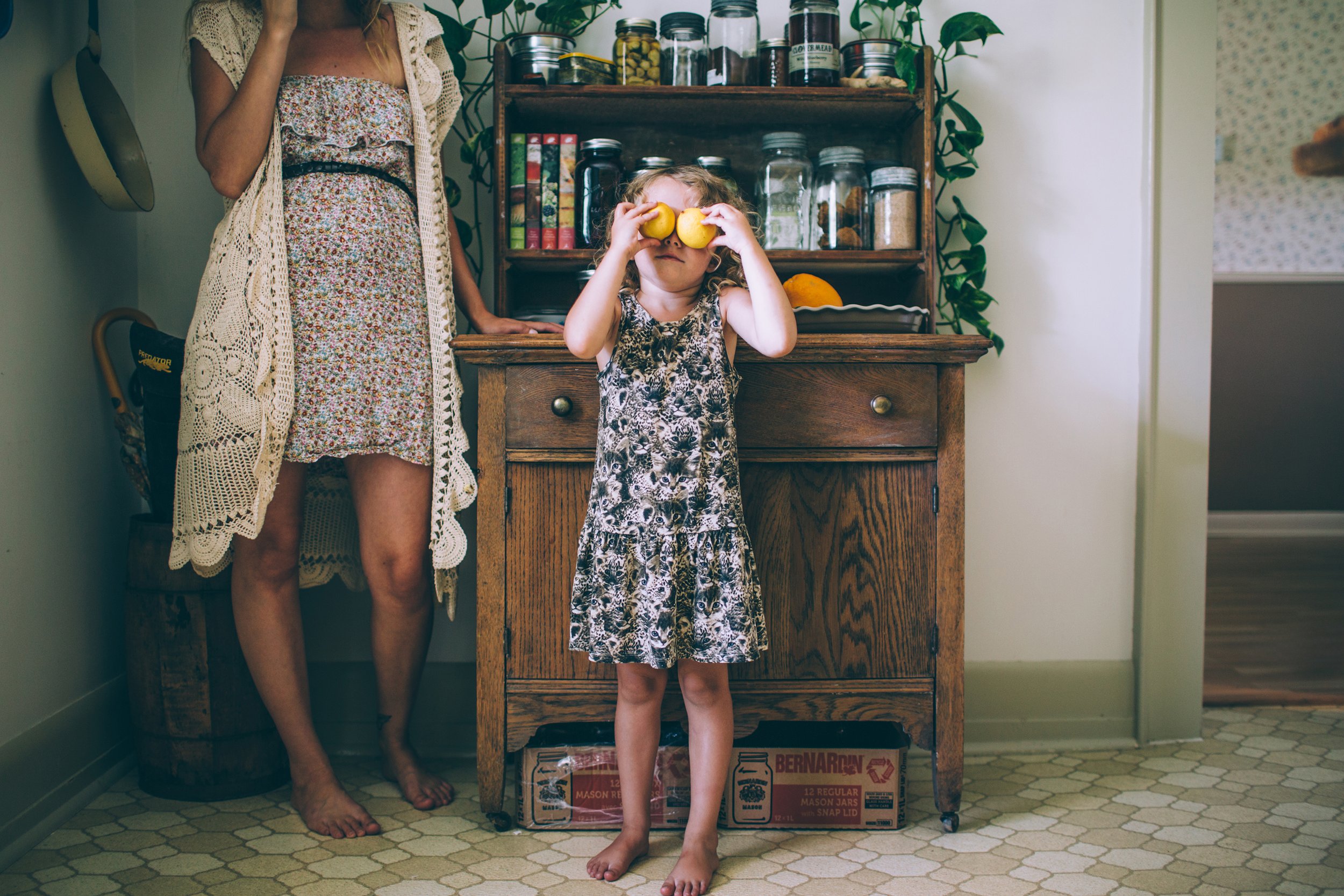 A girl playfully holding lemons in front of her eyes in a vintage-style kitchen, with a woman standing beside her.