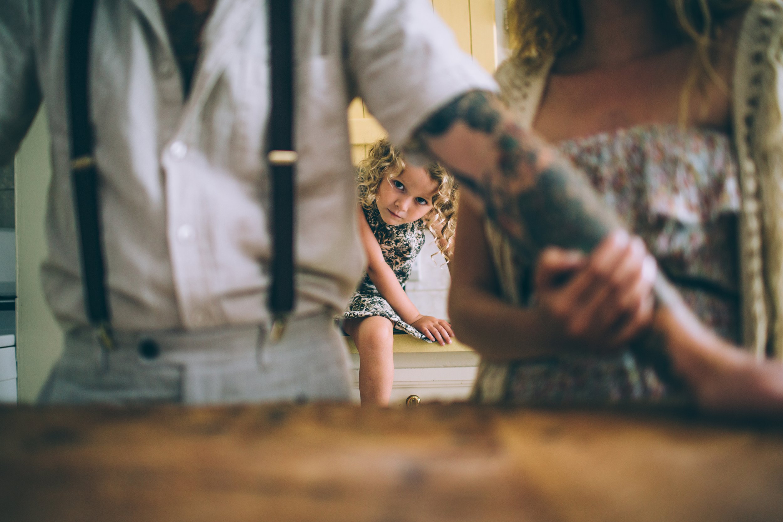 A young child sitting on a kitchen counter, looking curiously at the camera, while the focus is slightly blurred on the parents holding hands in the foreground.