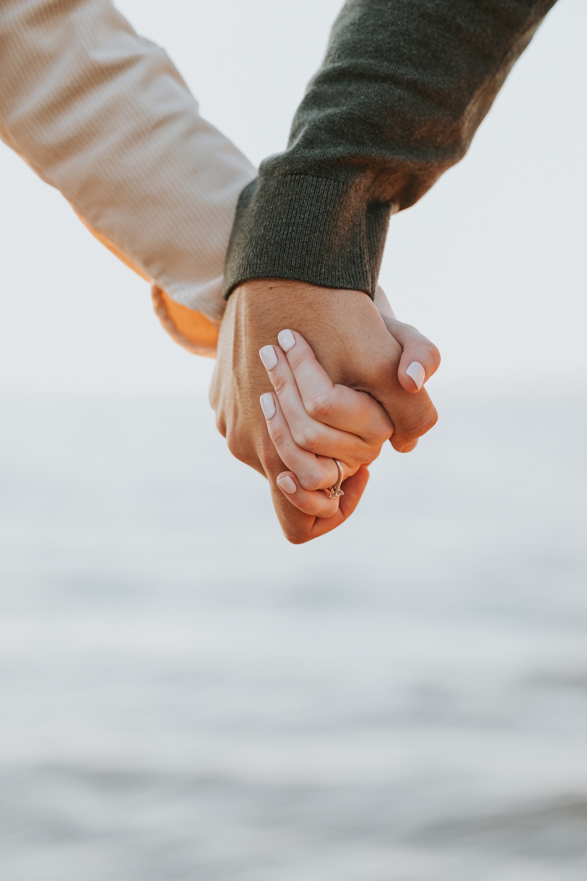 A close-up shot of a couple holding hands, showcasing a beautiful engagement ring. The intimate moment highlights their commitment and love, framed by the soft light of the setting sun over the ocean.