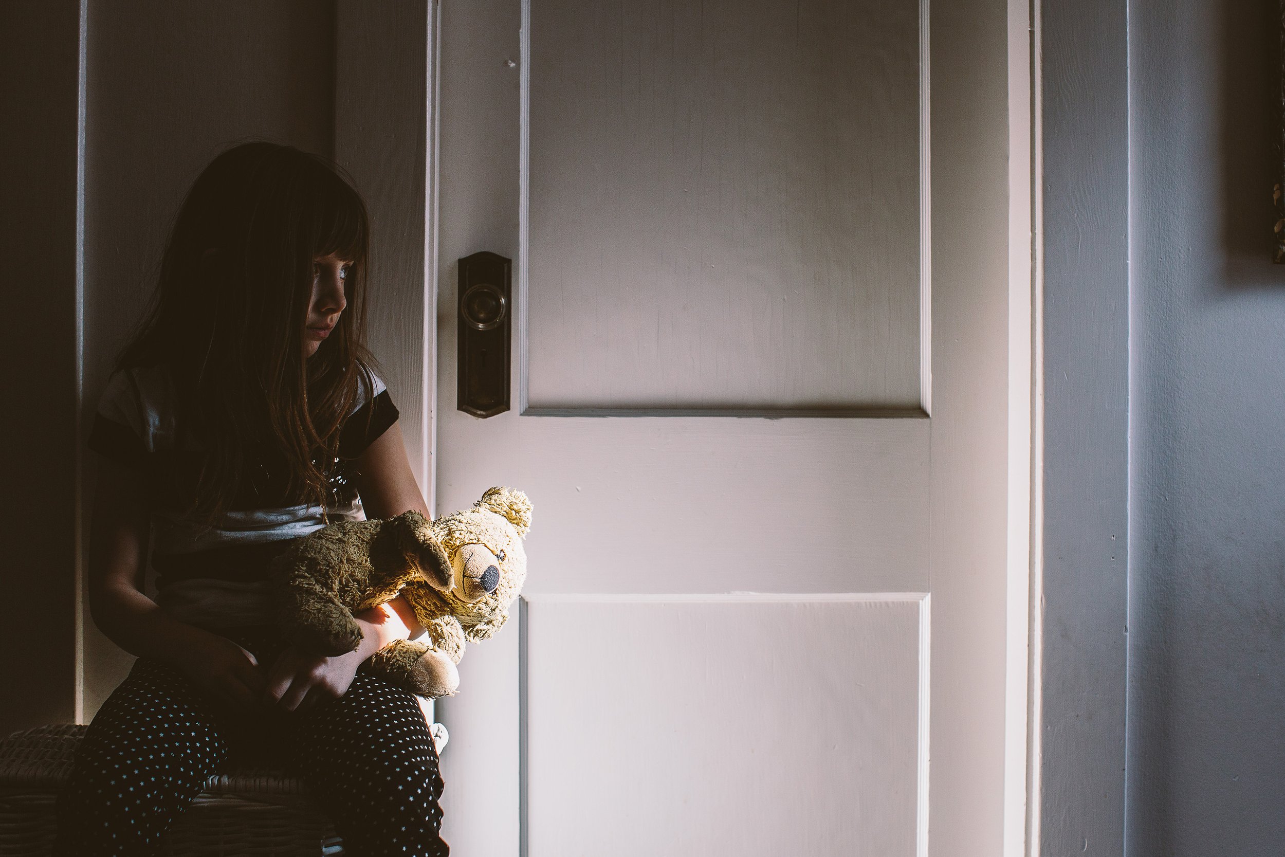 A child sitting in a doorway, holding a teddy bear, with soft light streaming in, creating a moment of quiet reflection.