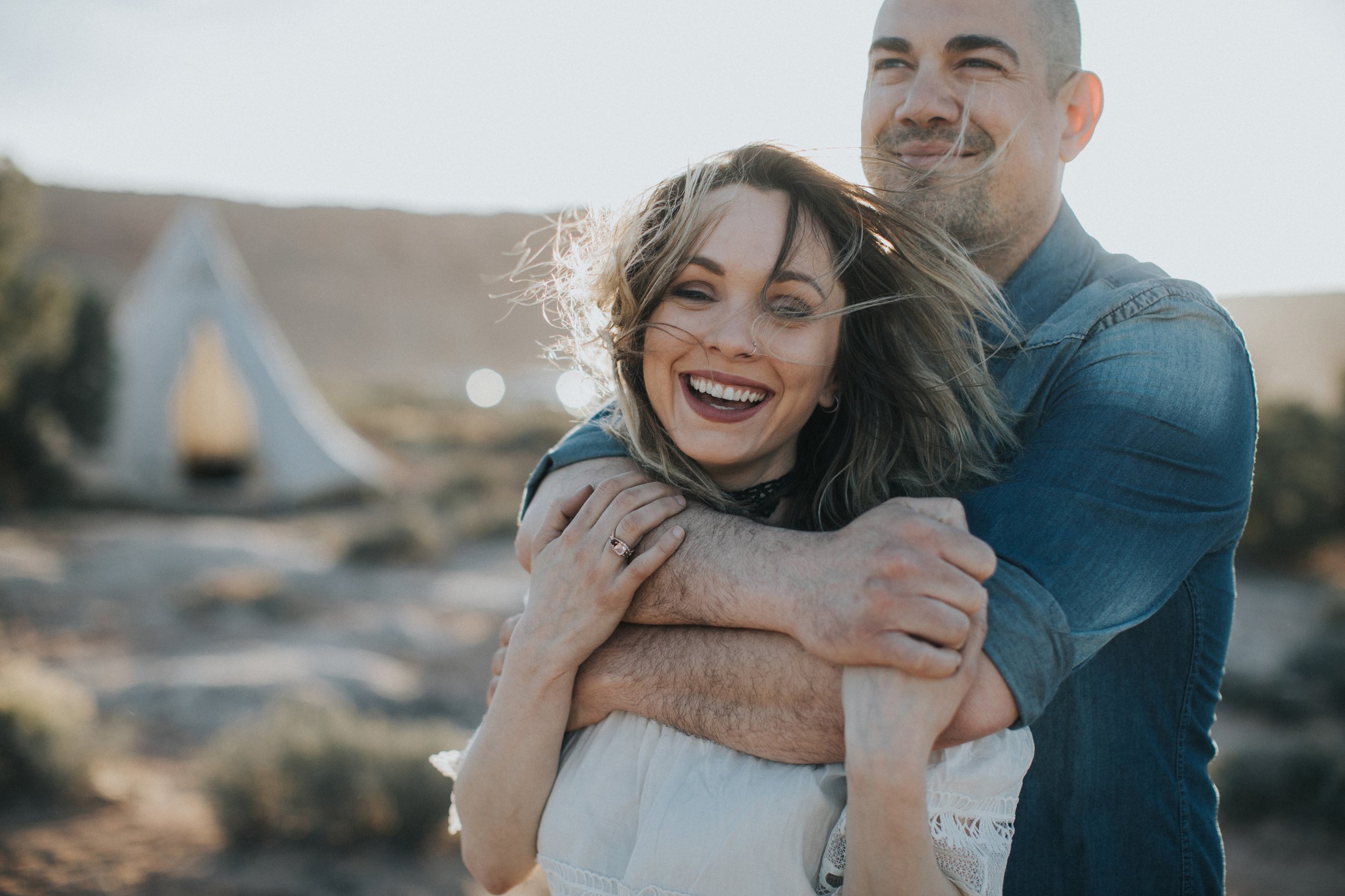 A couple laughing together in a windy desert landscape, capturing a joyful and carefree moment.