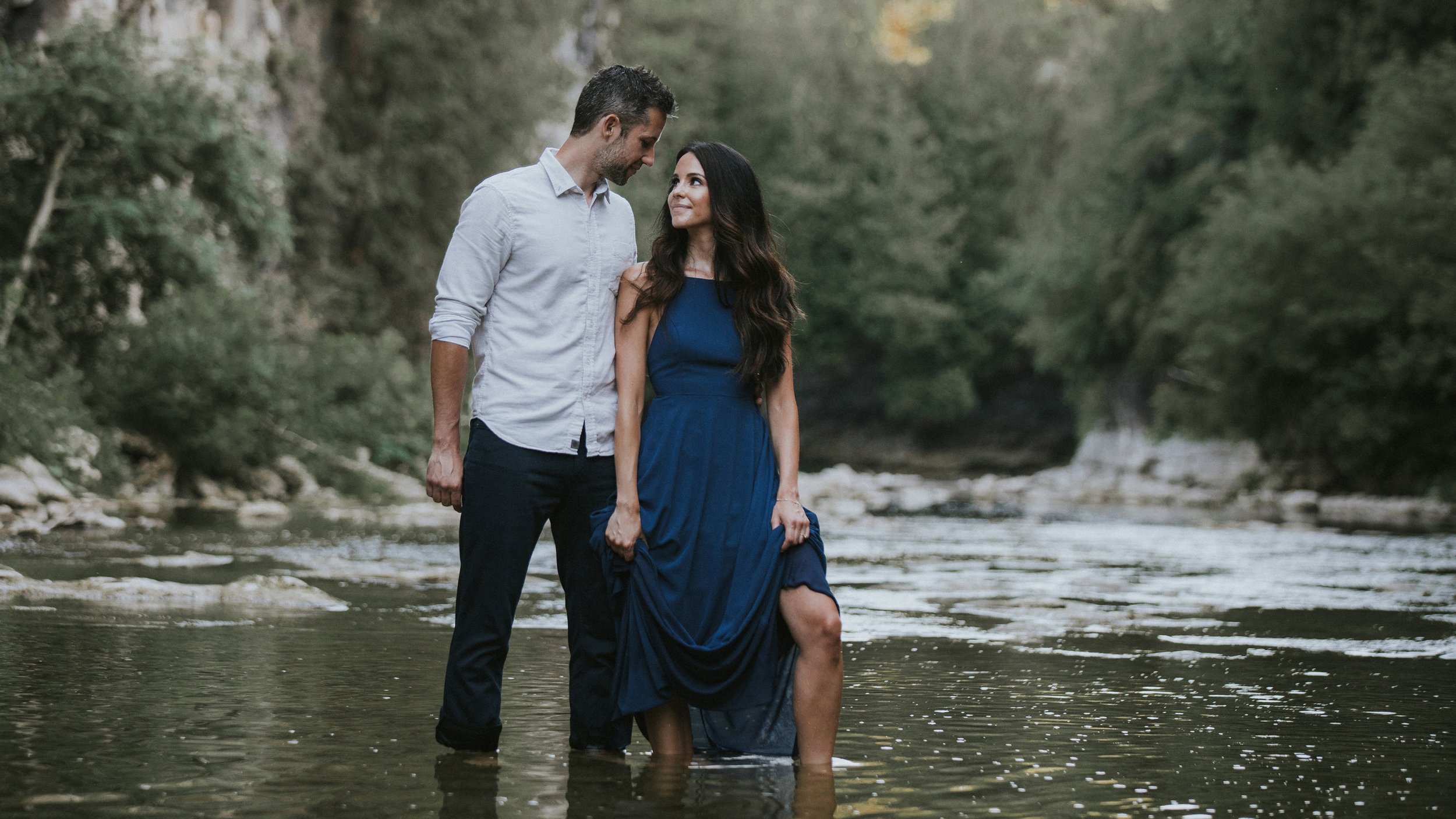 Couple standing in a river, gazing at each other lovingly, surrounded by lush greenery during their engagement session.
