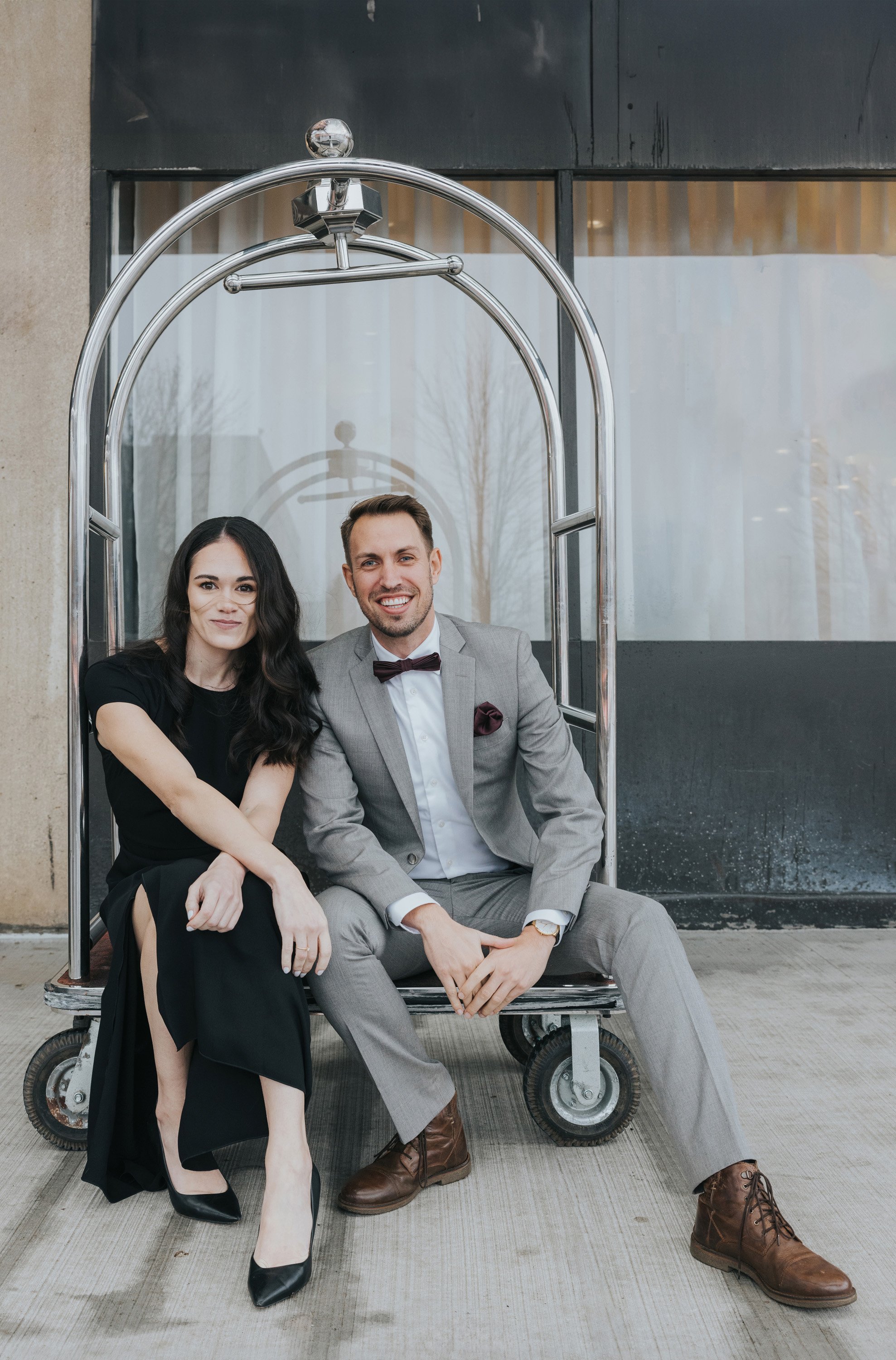 An elegant couple poses stylishly on a hotel bell cart, dressed in formal attire. The woman’s black dress and the man’s gray suit add a touch of glamour and sophistication to the scene.