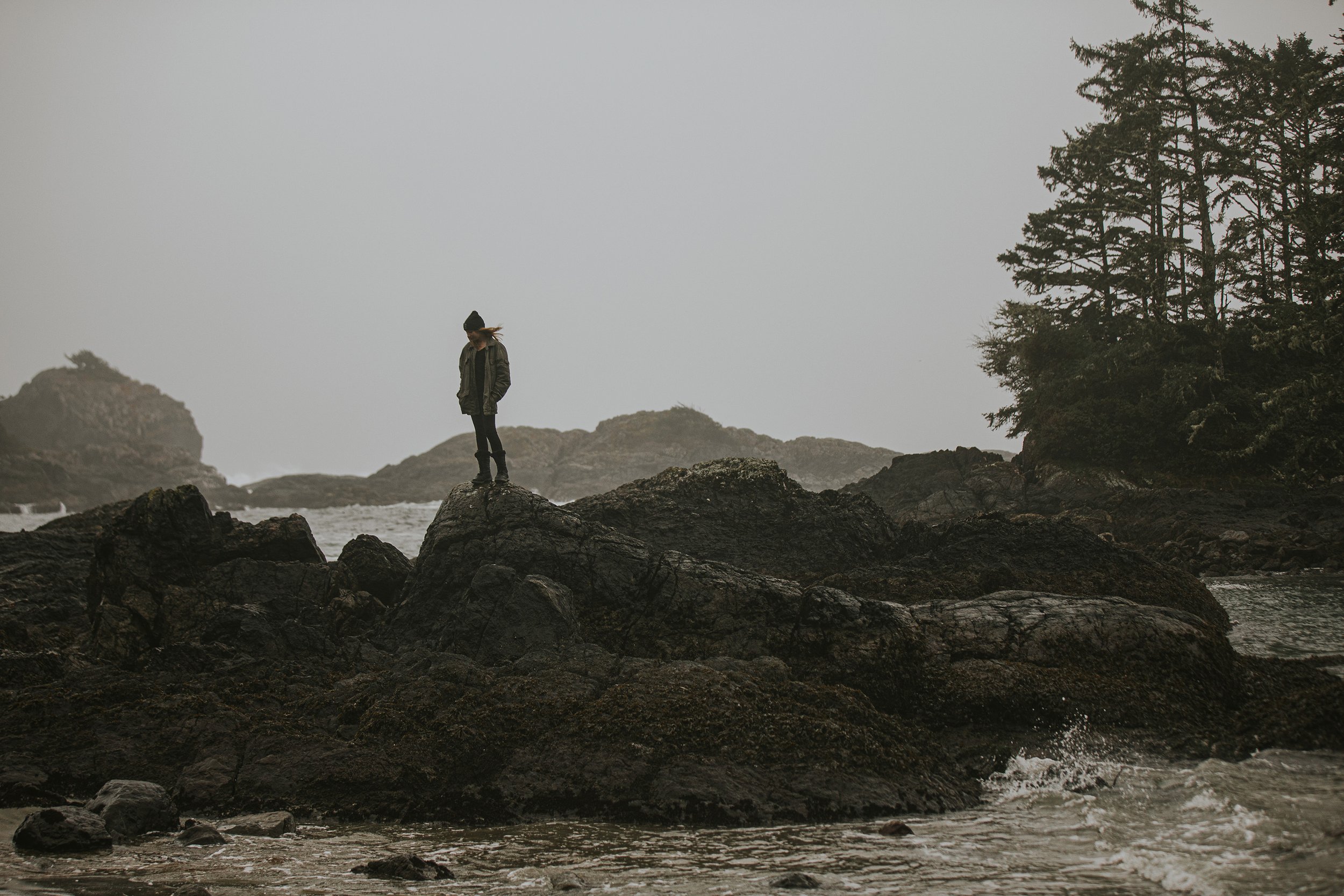 A young girl standing alone on a rocky shoreline, with a foggy coast providing a moody backdrop.