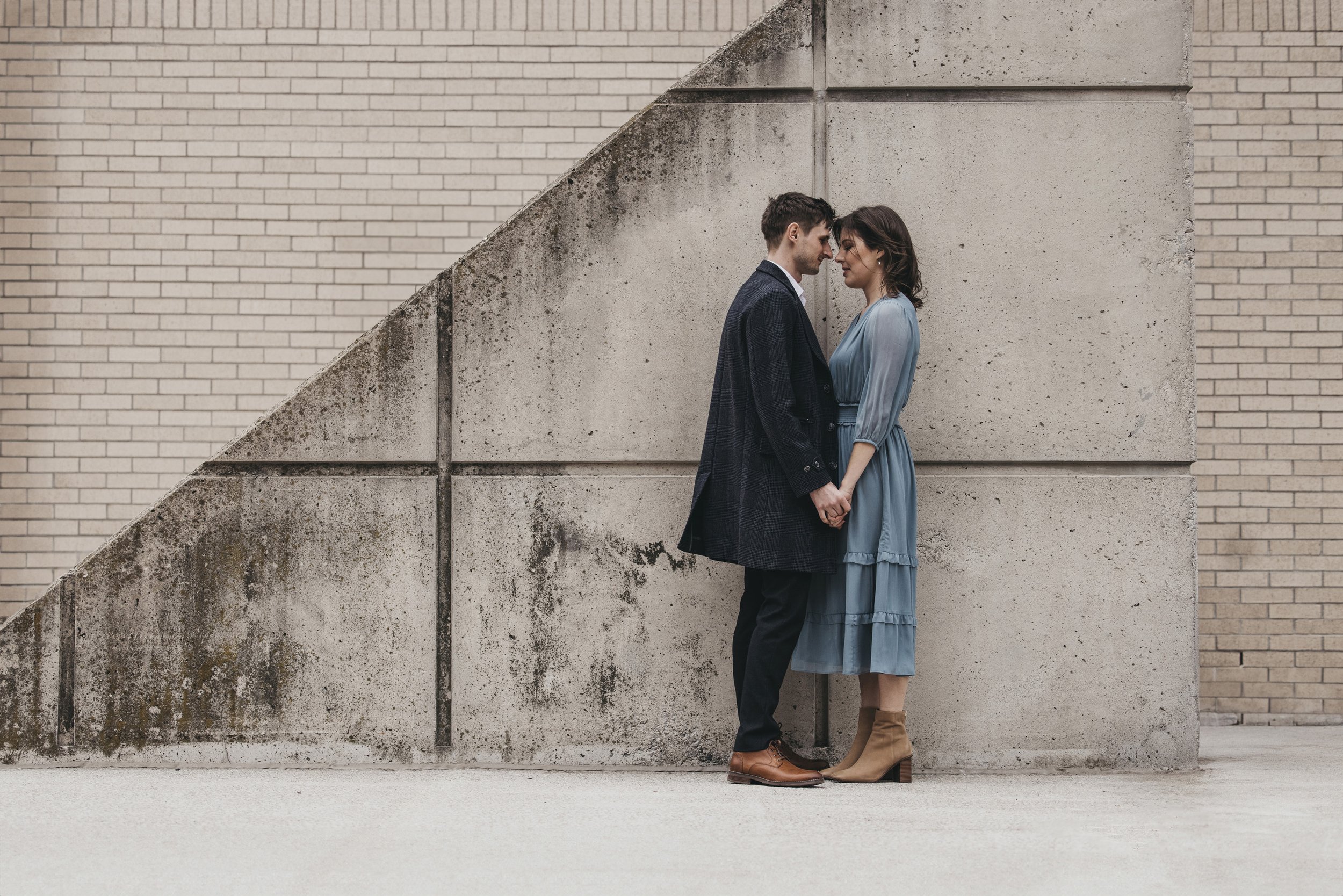 A couple holds hands while standing against a concrete wall in an urban setting, emphasizing their connection.