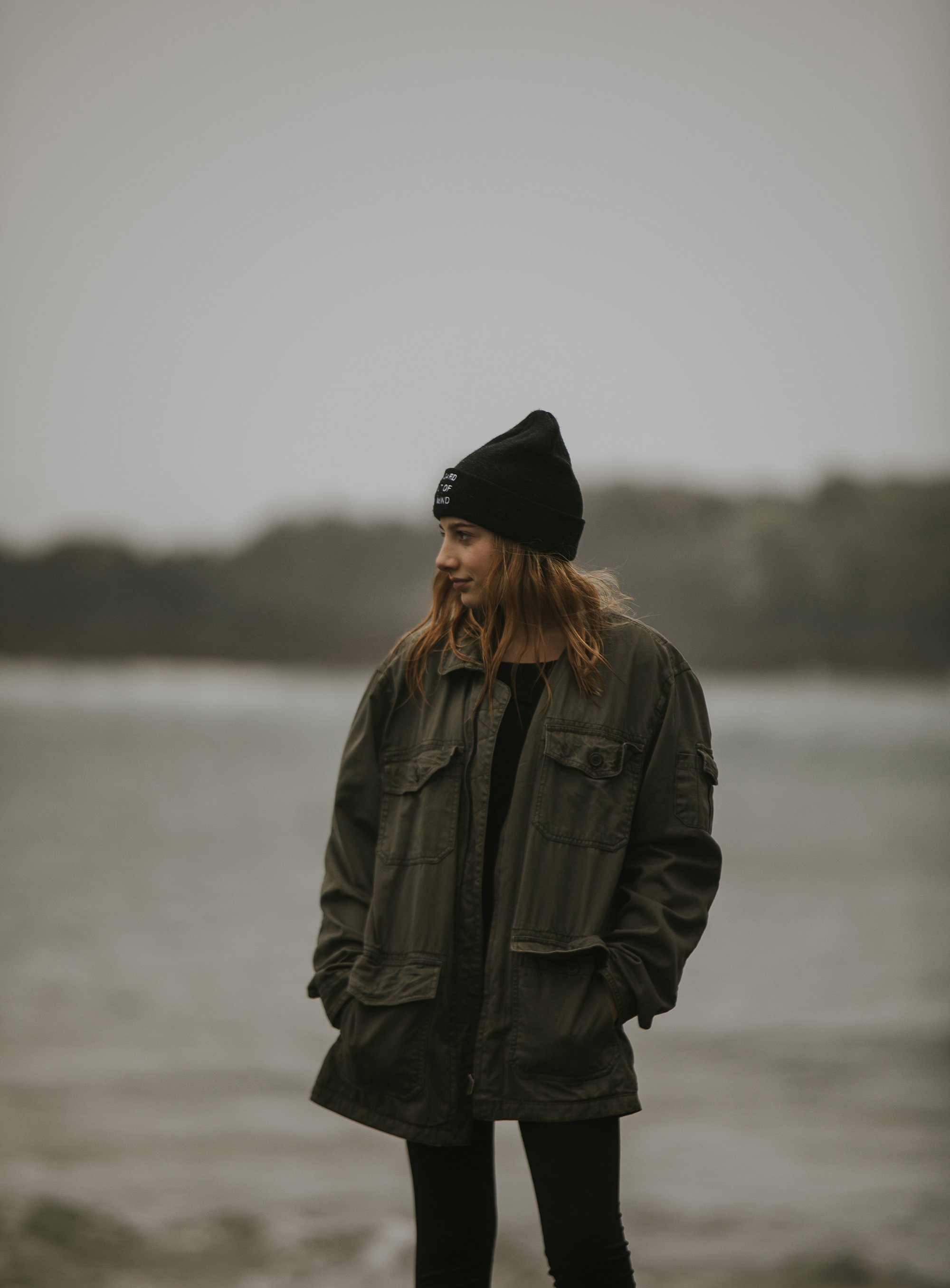 Teen Girl Outdoor Jacket Moody Beach Portrait: A teen girl wearing a beanie and a jacket stands by the beach, with a moody, overcast background.
