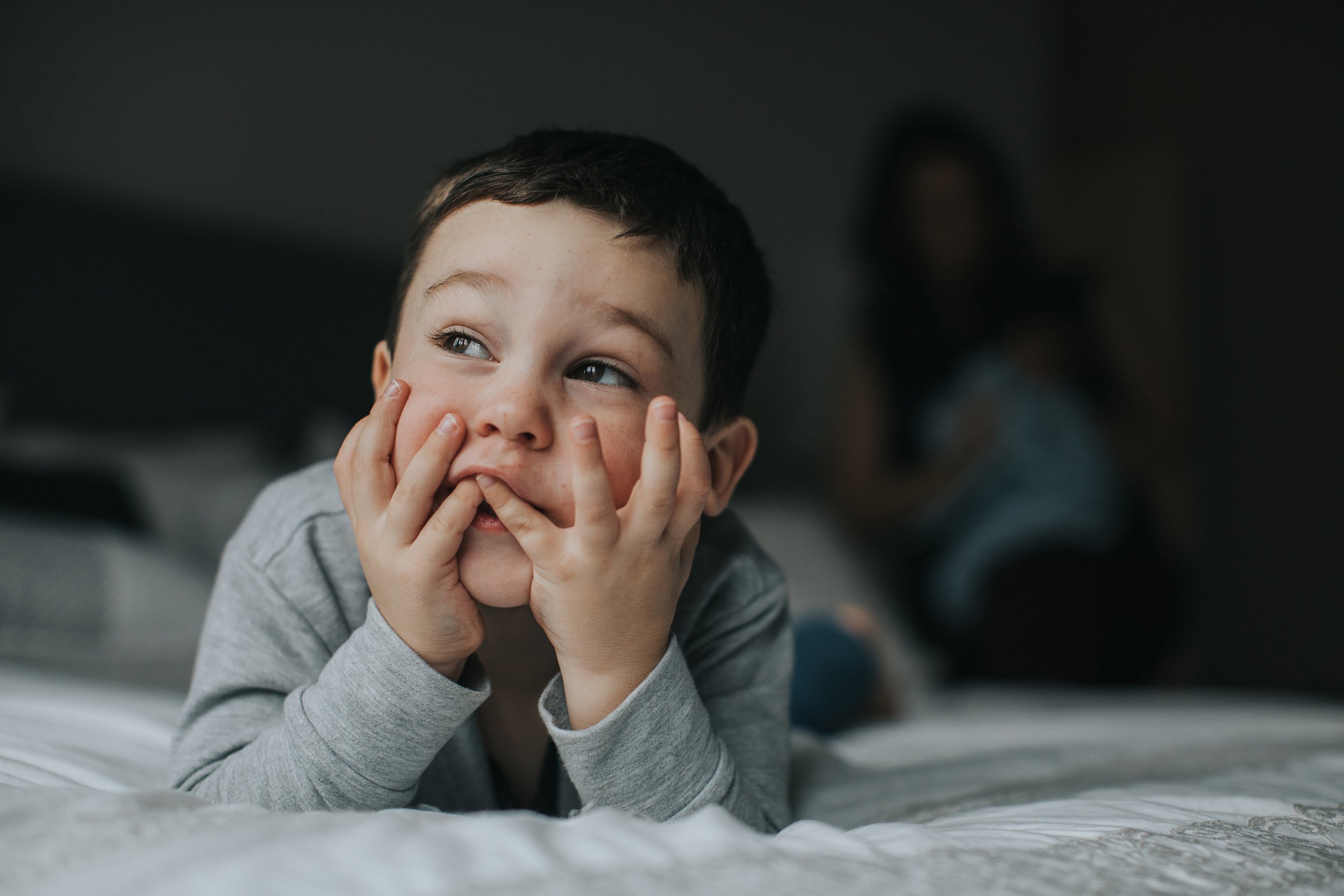 A playful moment of a young boy lying on a bed with a thoughtful expression, resting his face on his hands, captured in a cozy indoor setting.