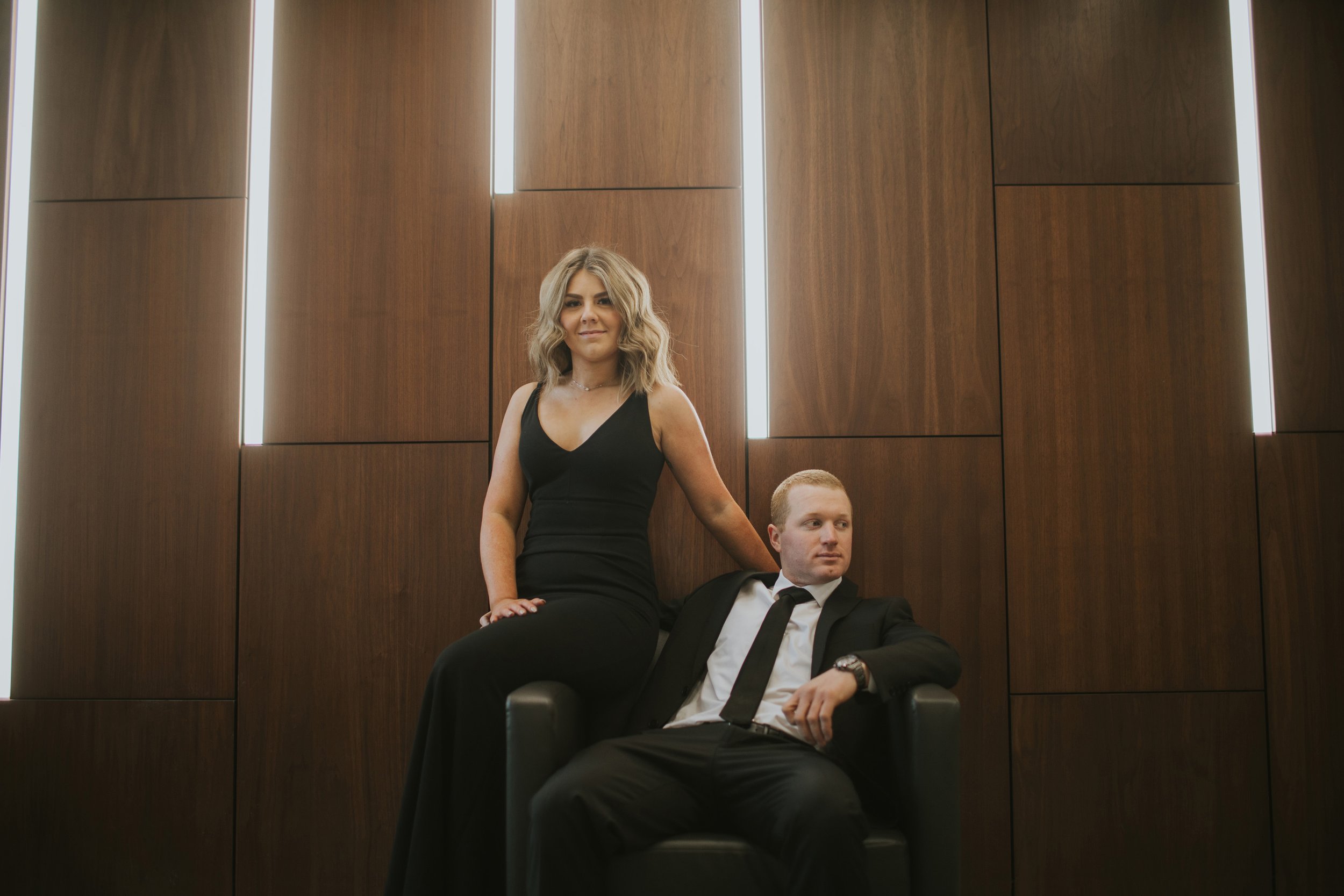 A stylish couple posing indoors during their engagement portrait session, dressed in formal attire against a modern backdrop.
