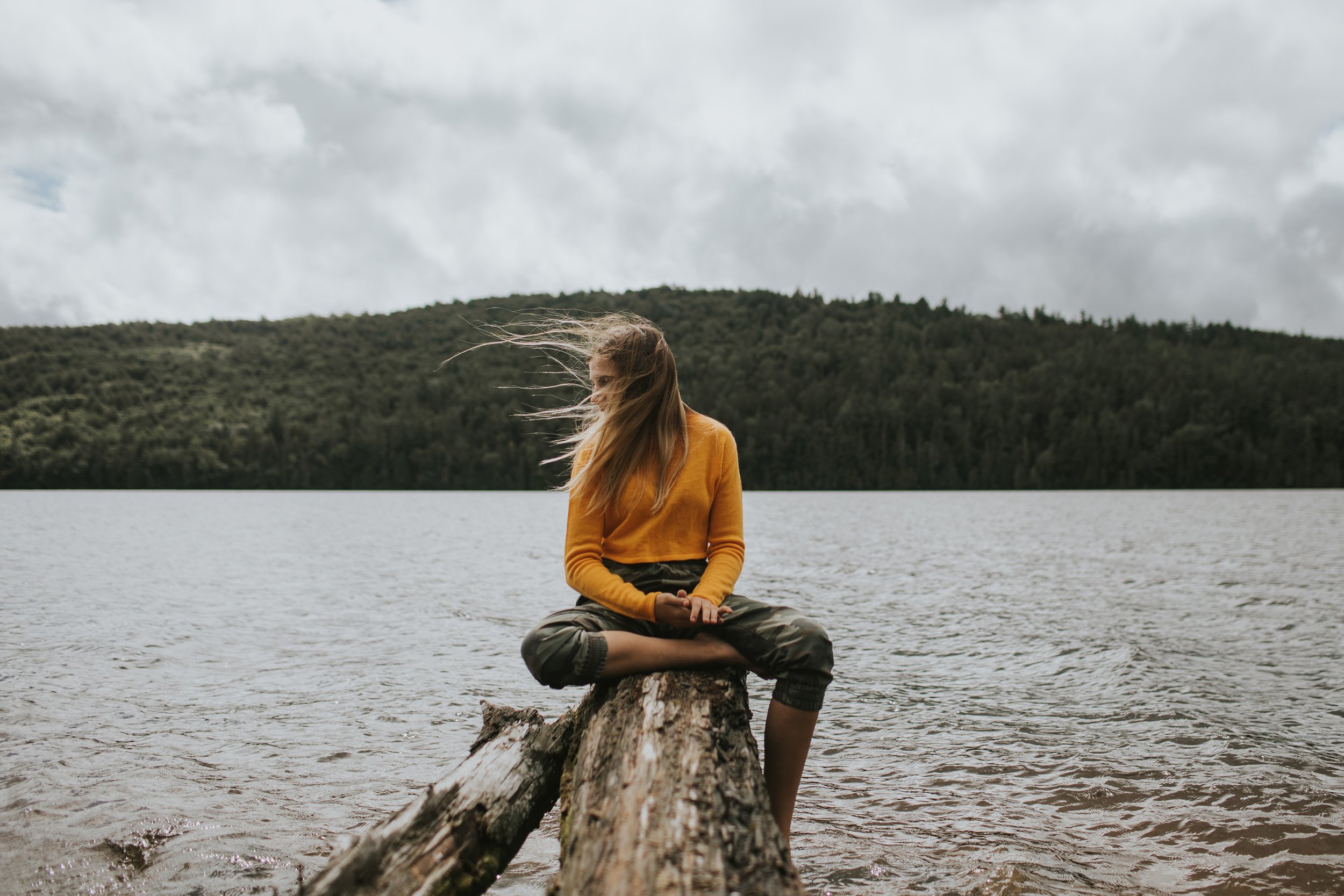 Woman Sitting Log Lake Nature Wind Adventure London Ontario: A woman in an orange sweater sitting cross-legged on a log by a lake, with her hair blowing in the wind.