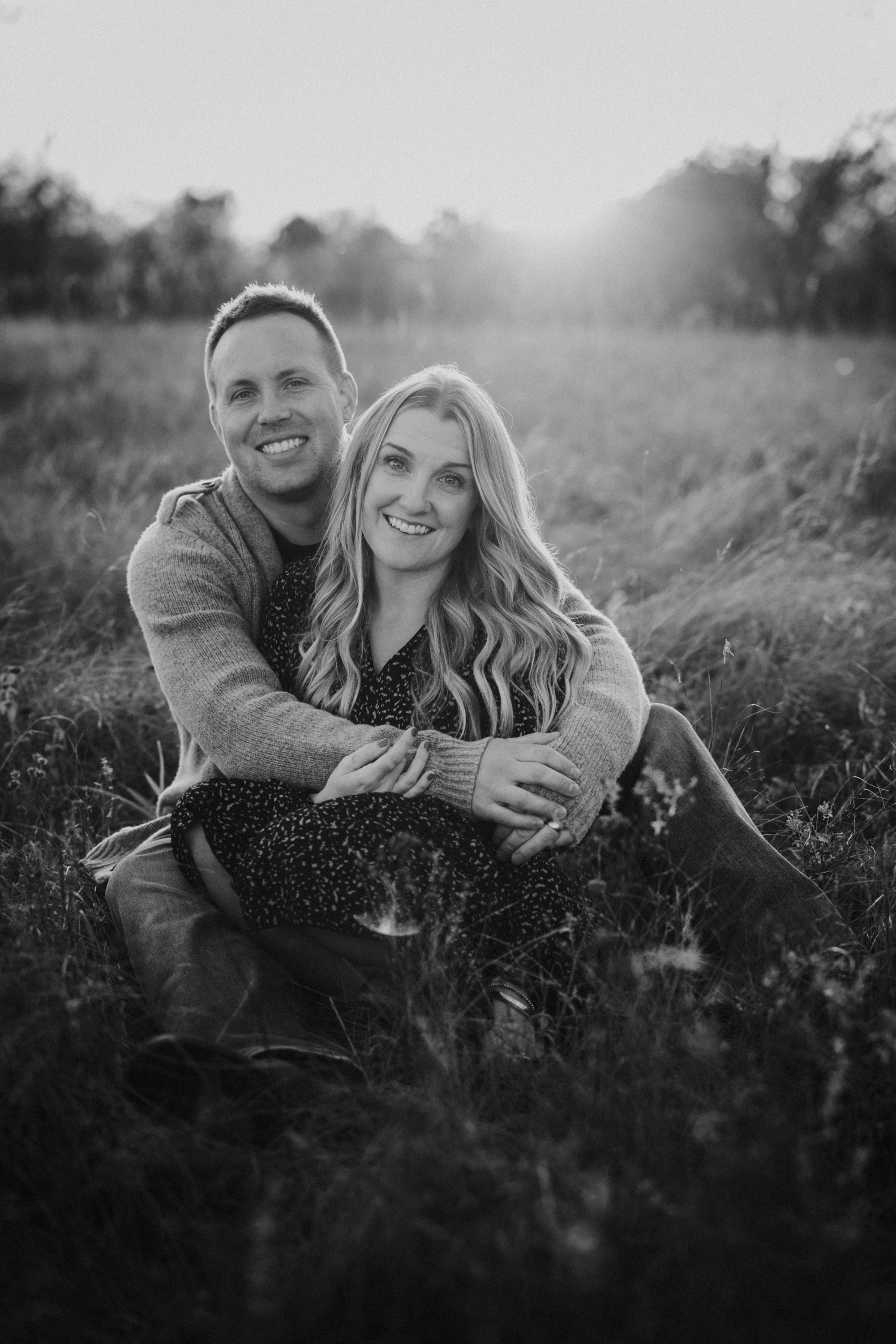 A black-and-white photo of a couple embracing in a field at sunset, capturing an intimate moment.