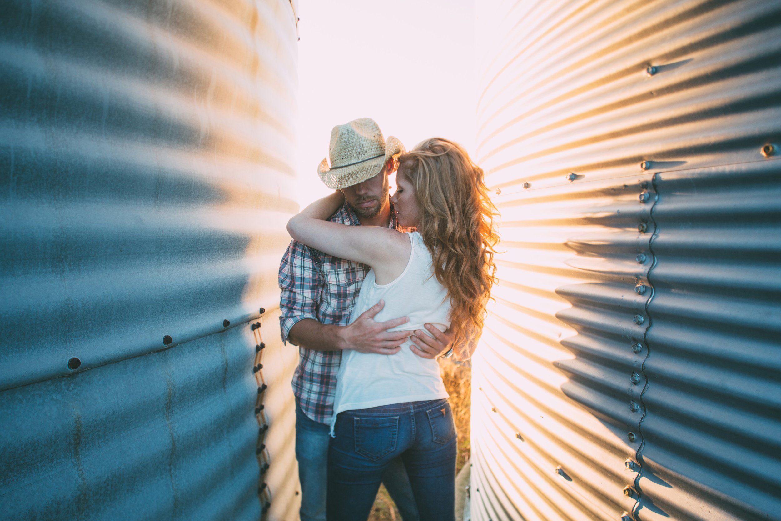 A couple embraces warmly between two farm silos, with the setting sun casting a golden glow on their figures. The man wears a plaid shirt and cowboy hat, while the woman’s hair catches the light, creating a romantic and rustic atmosphere.