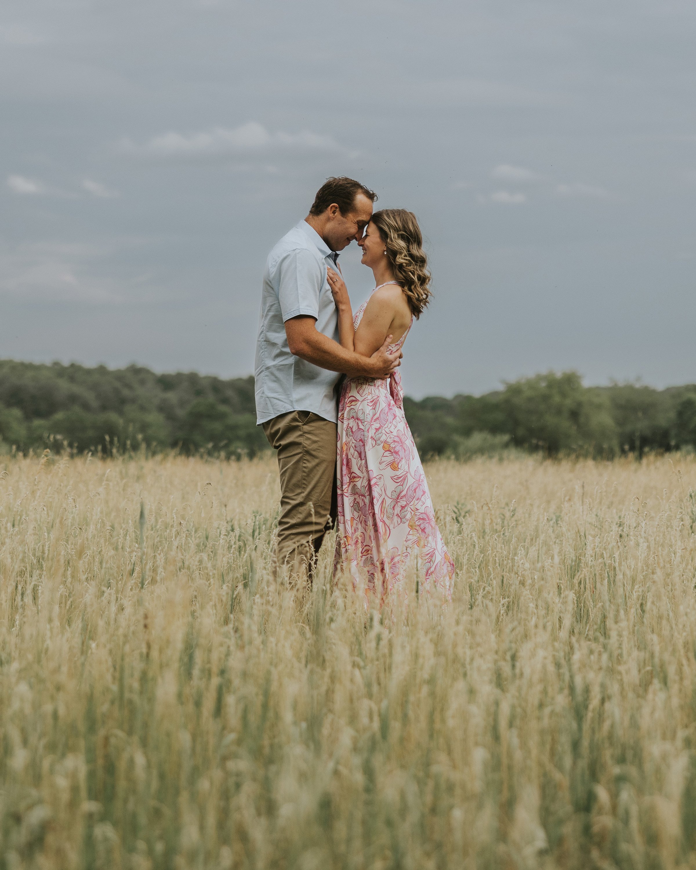 A couple stands close together in a sunlit meadow, surrounded by tall grass, enjoying a tender moment during golden hour.