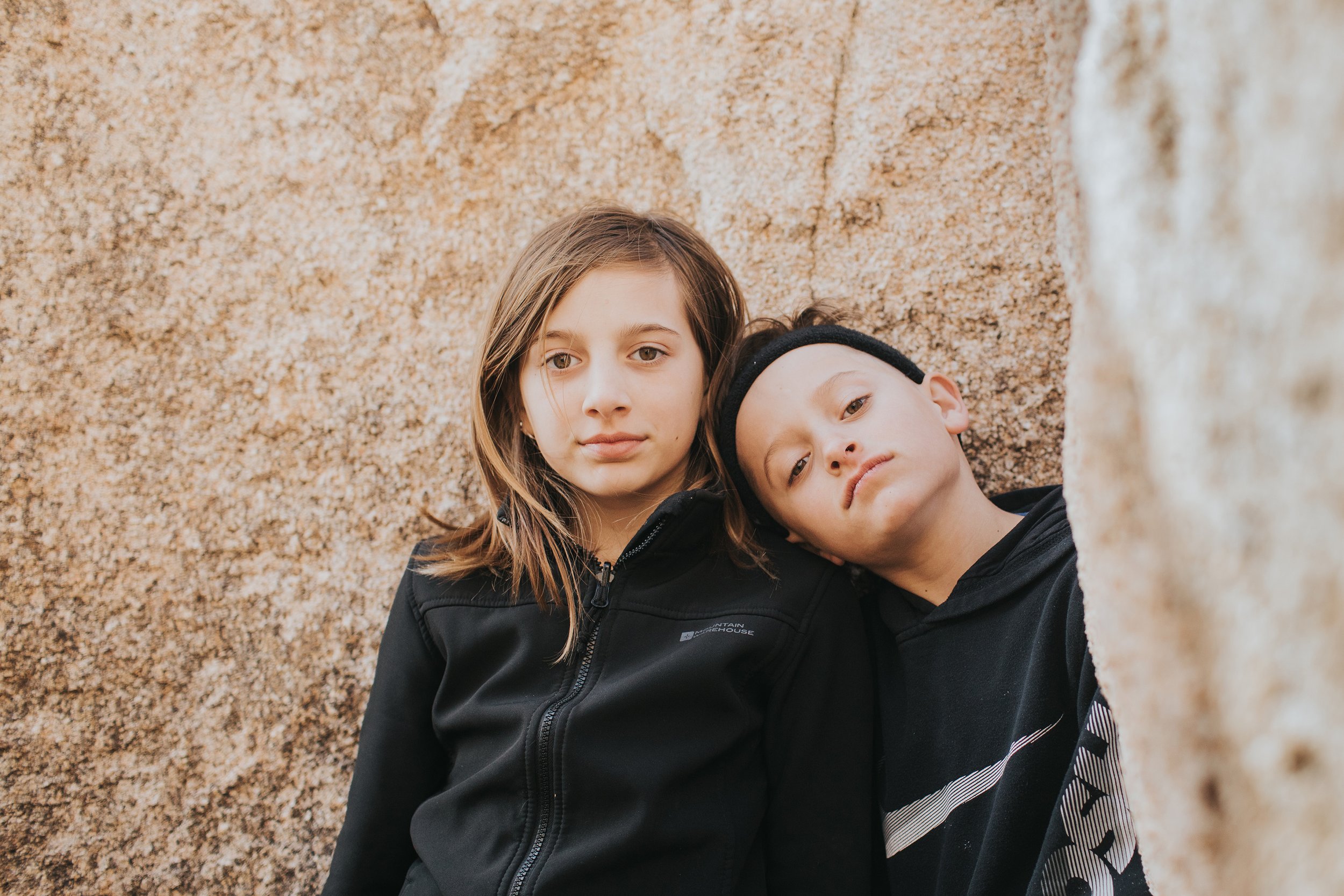 Close-up of a brother and sister leaning against a large rock formation, looking calm and connected in a natural setting.