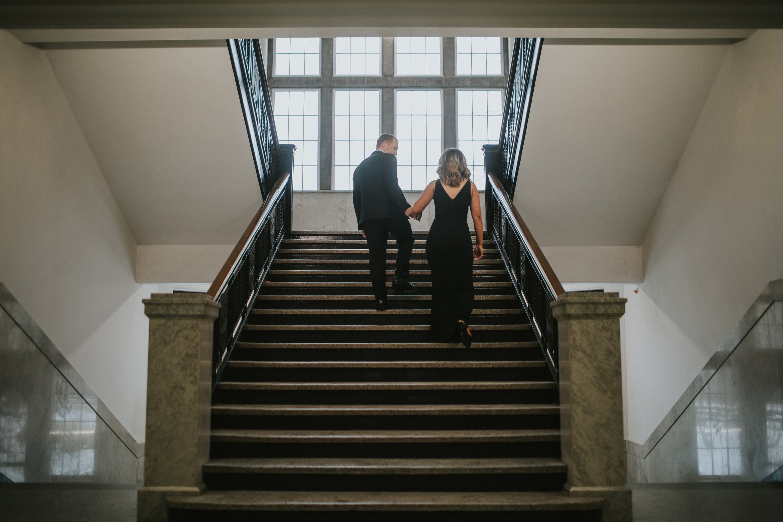 Elegant couple walking up a grand staircase, hand-in-hand, during their engagement session in a classic indoor setting.