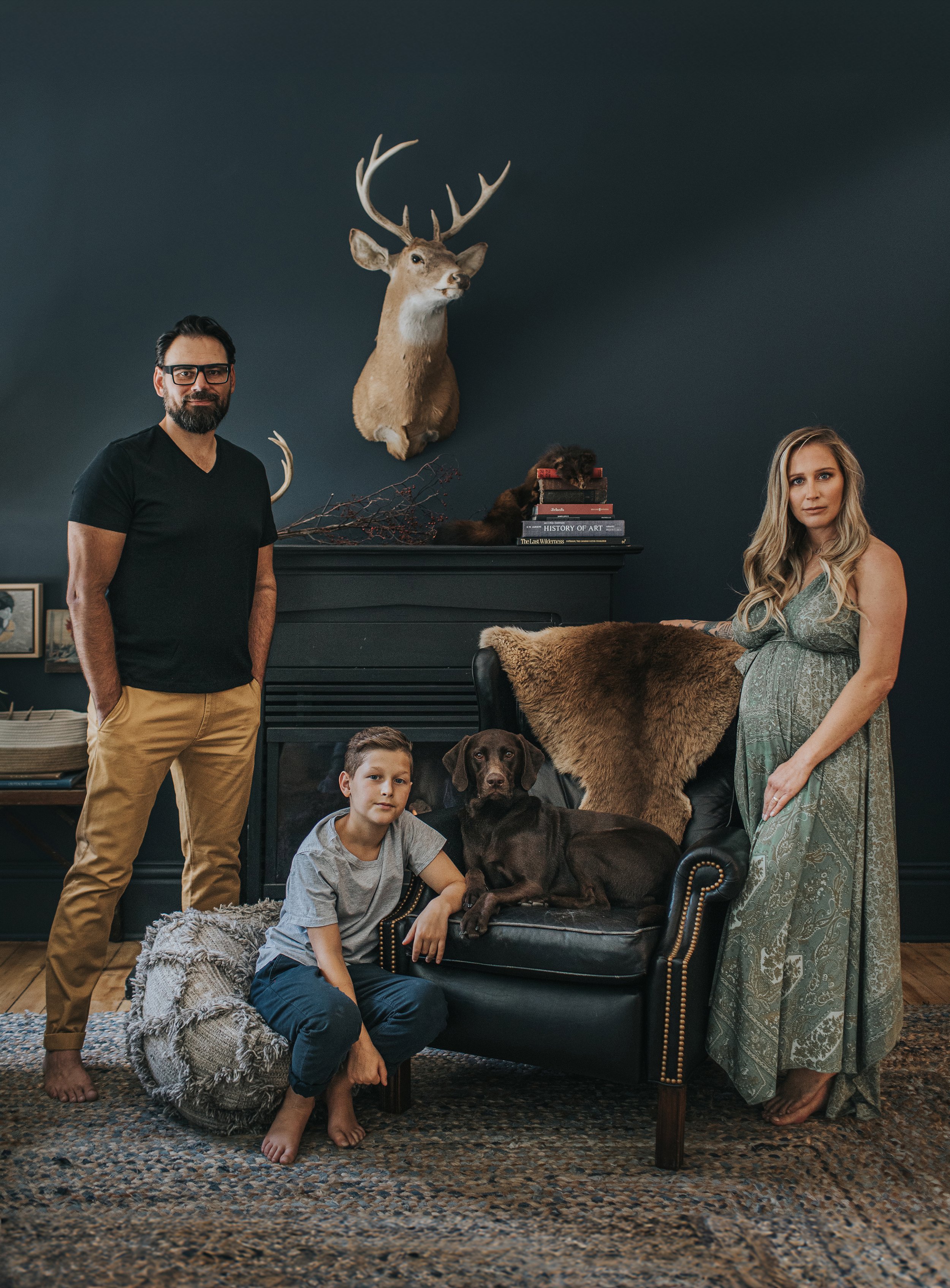 A formal family portrait in a classic living room, featuring parents, their son, and their dog, with dark walls and a mounted deer head in the background.