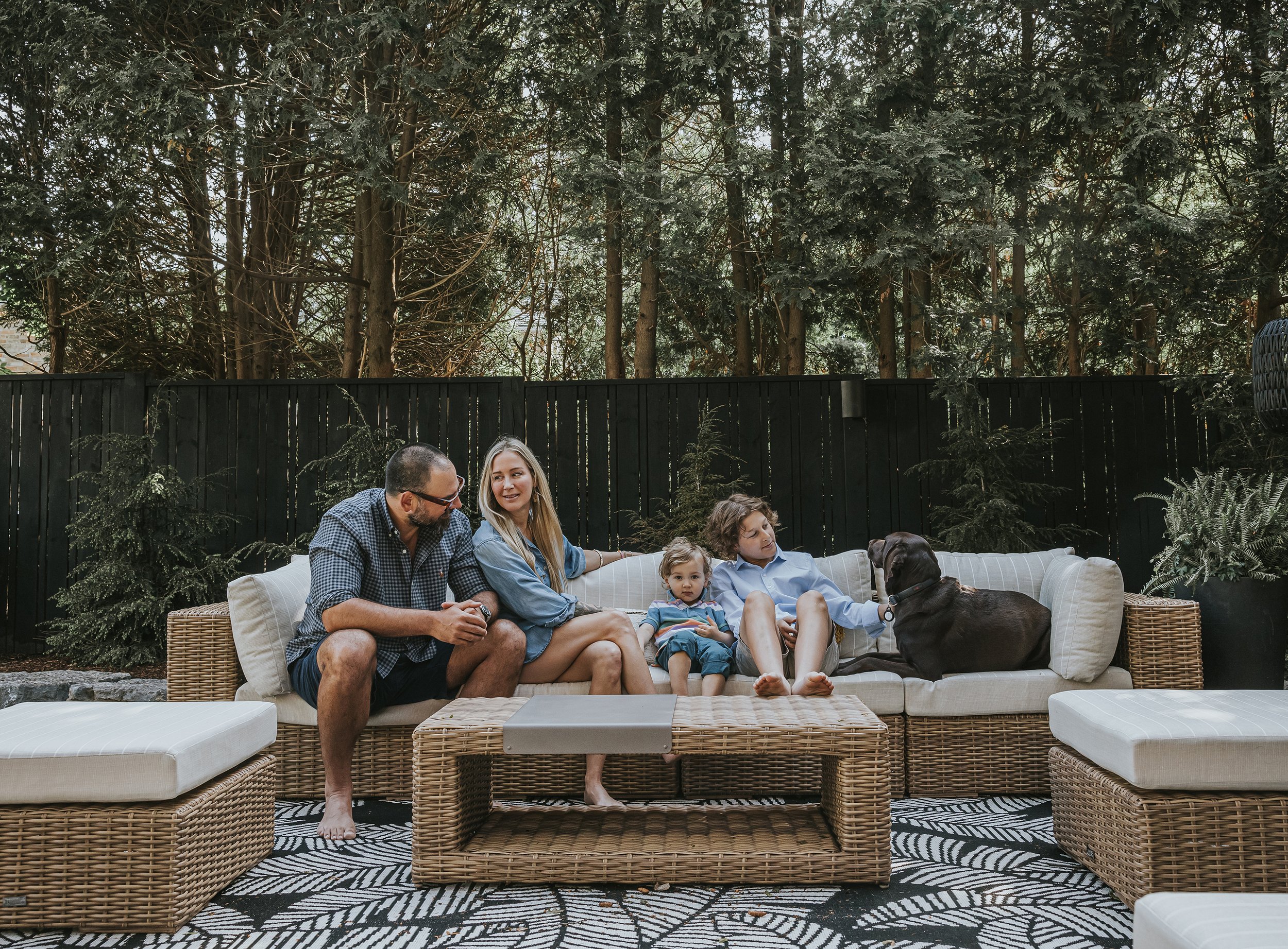 A relaxed family moment captured outdoors on a wicker sofa, with trees providing a natural backdrop, featuring parents and two young children with their dog.