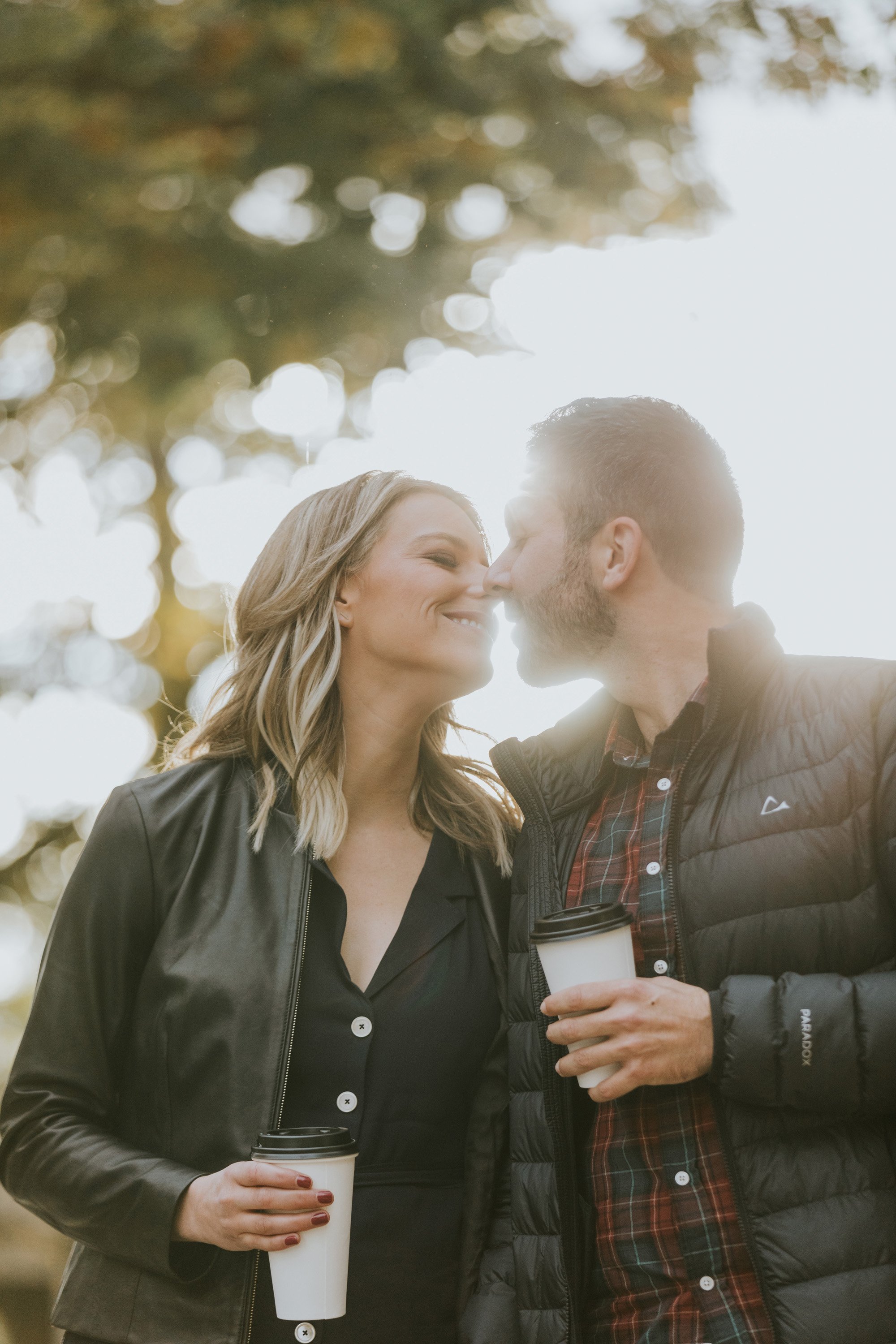 A couple shares a tender kiss while holding coffee cups during an autumn engagement session. The sun shines brightly behind them, creating a warm and intimate moment.
