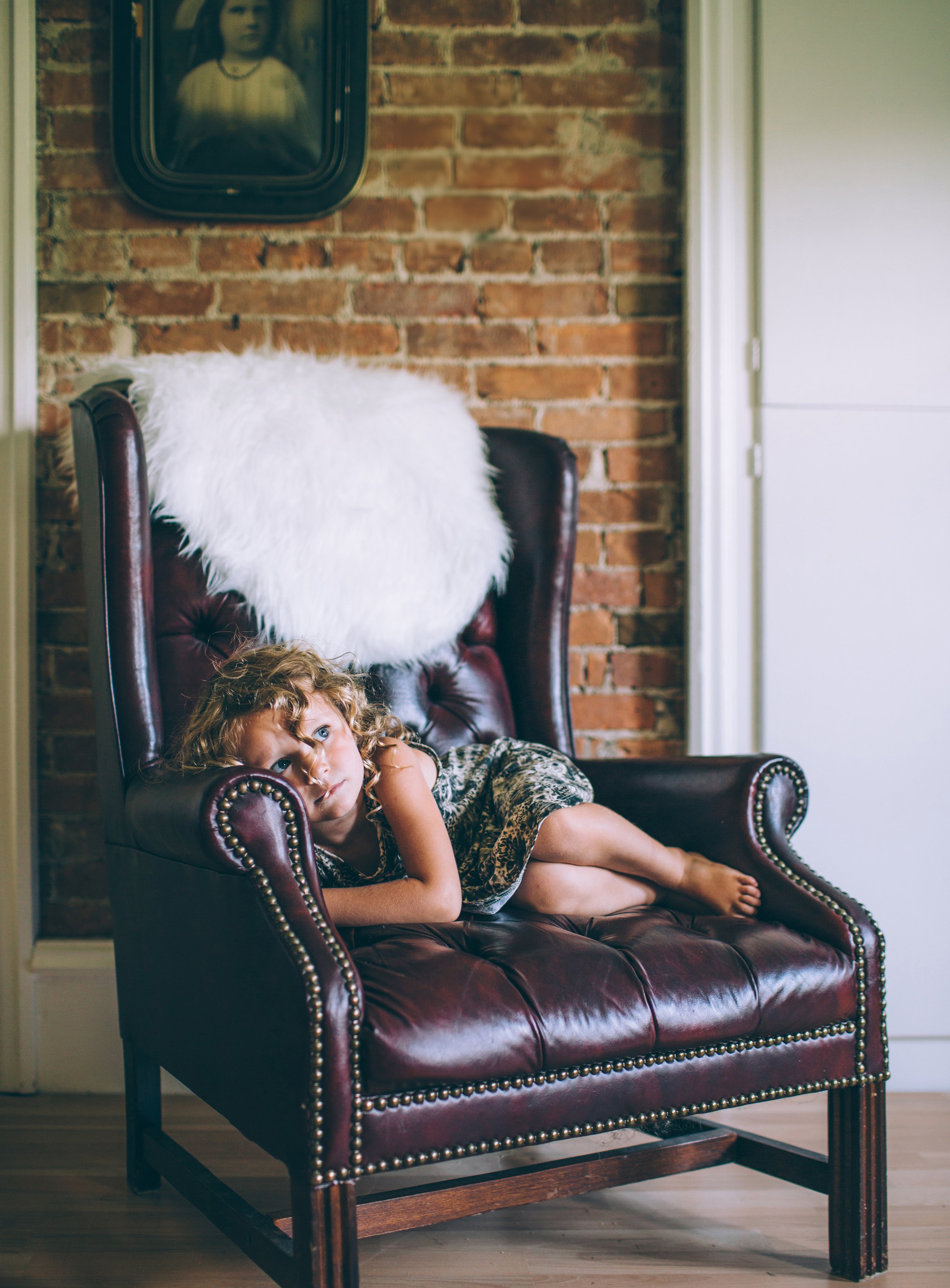 A young child lounging on a vintage leather chair in a room with exposed brick walls.