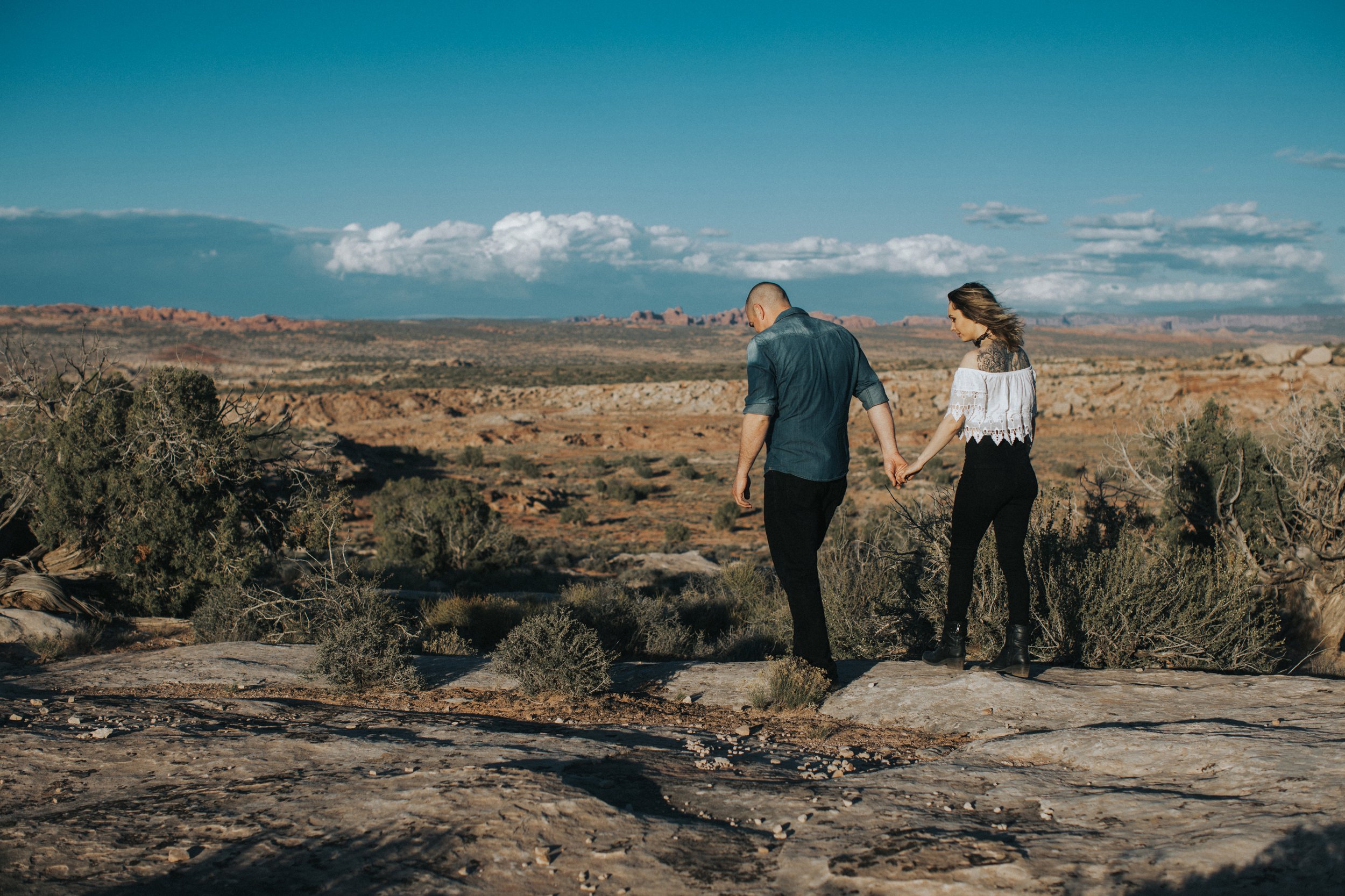 A couple holding hands while walking through a desert landscape during their engagement session.