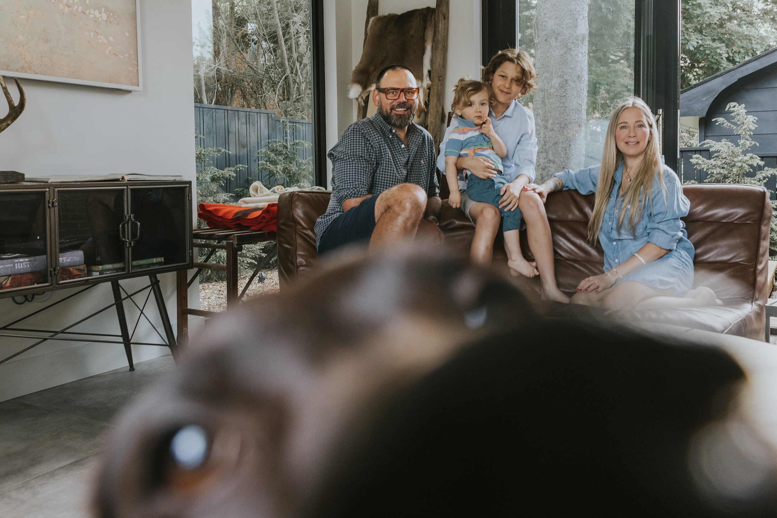 A family sitting on a leather couch in a modern living room, with their pet dog playfully photobombing the scene in the foreground.