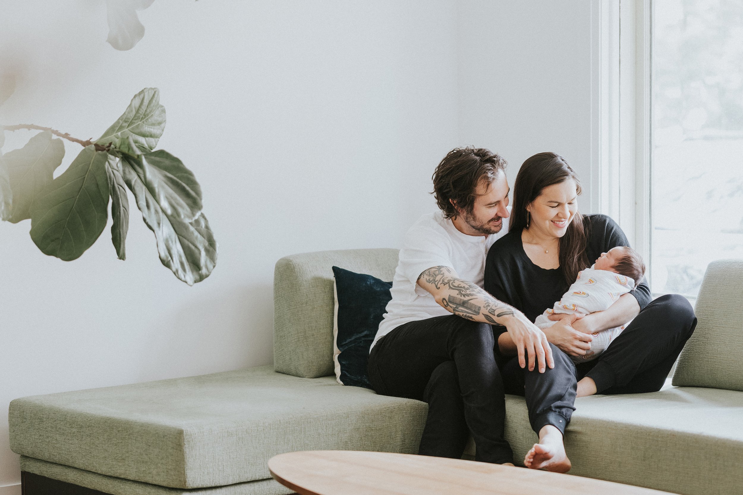 A candid moment of new parents sitting on a couch, lovingly holding their newborn baby in a bright, airy living room.
