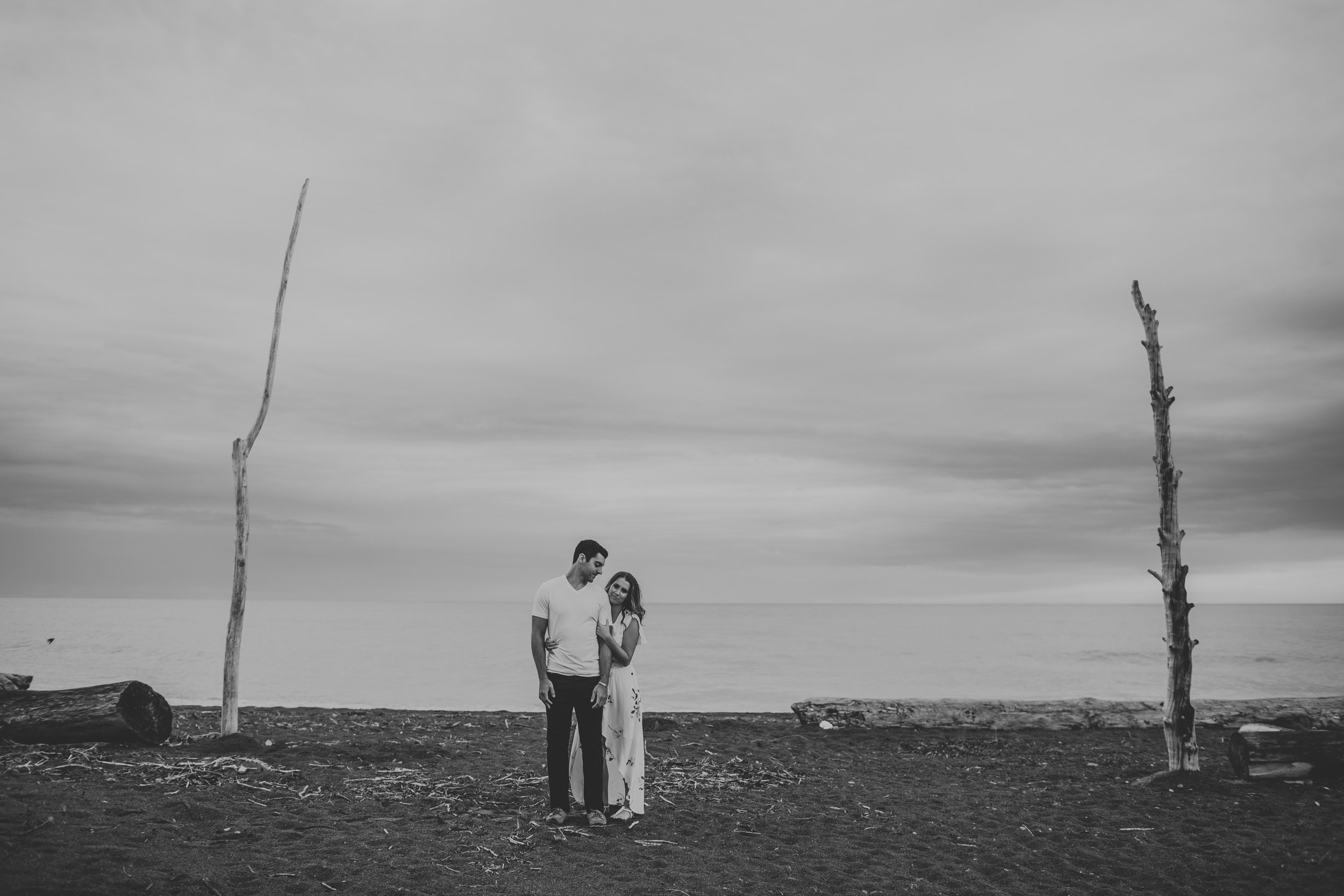 A minimalist black-and-white photograph of a couple standing on a beach near driftwood. The image captures a quiet, intimate moment in a serene natural setting.