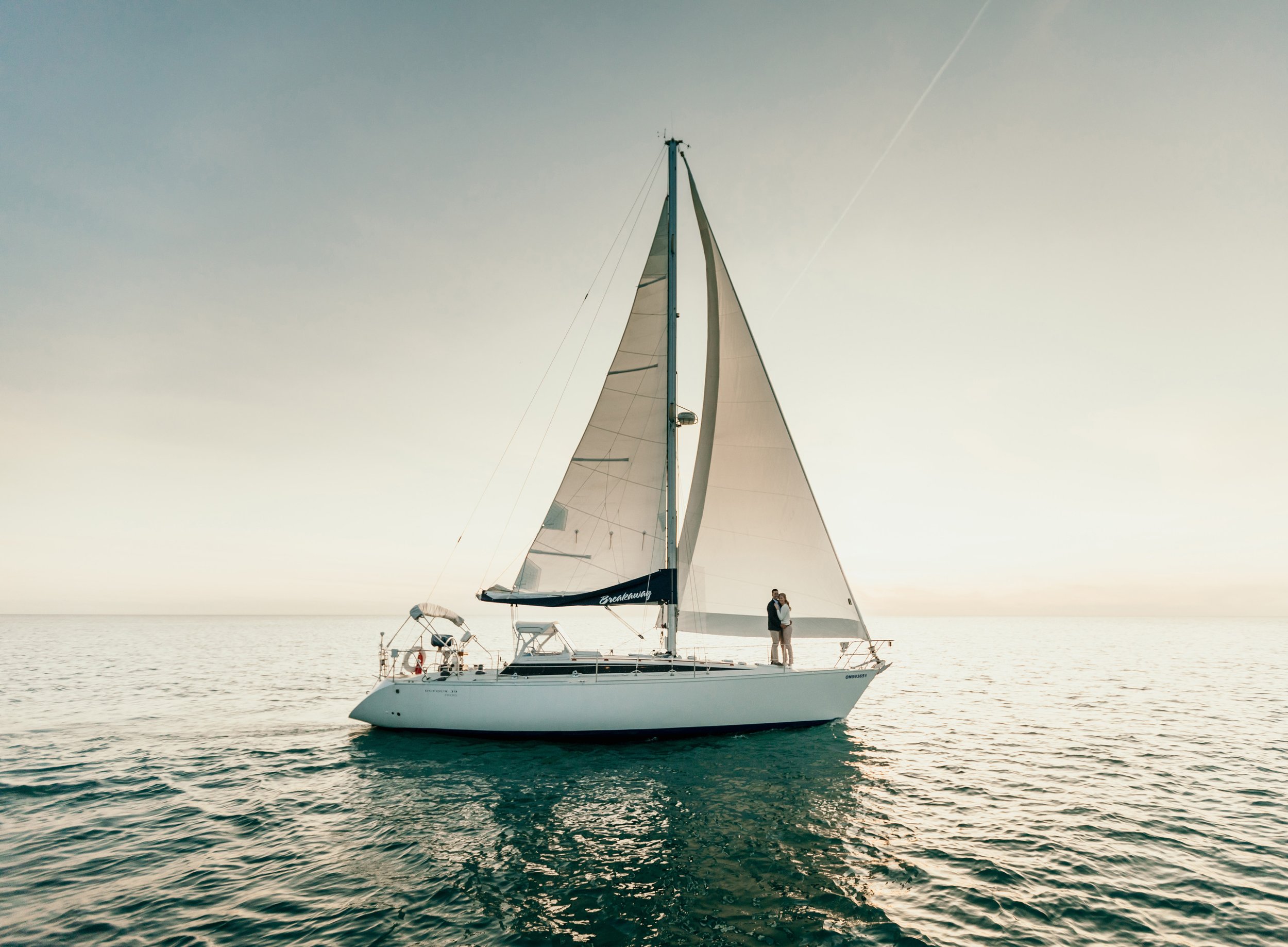 A couple standing on a luxurious yacht at sunset on a serene lake in Ontario, embracing as the sailboat glides through calm waters.