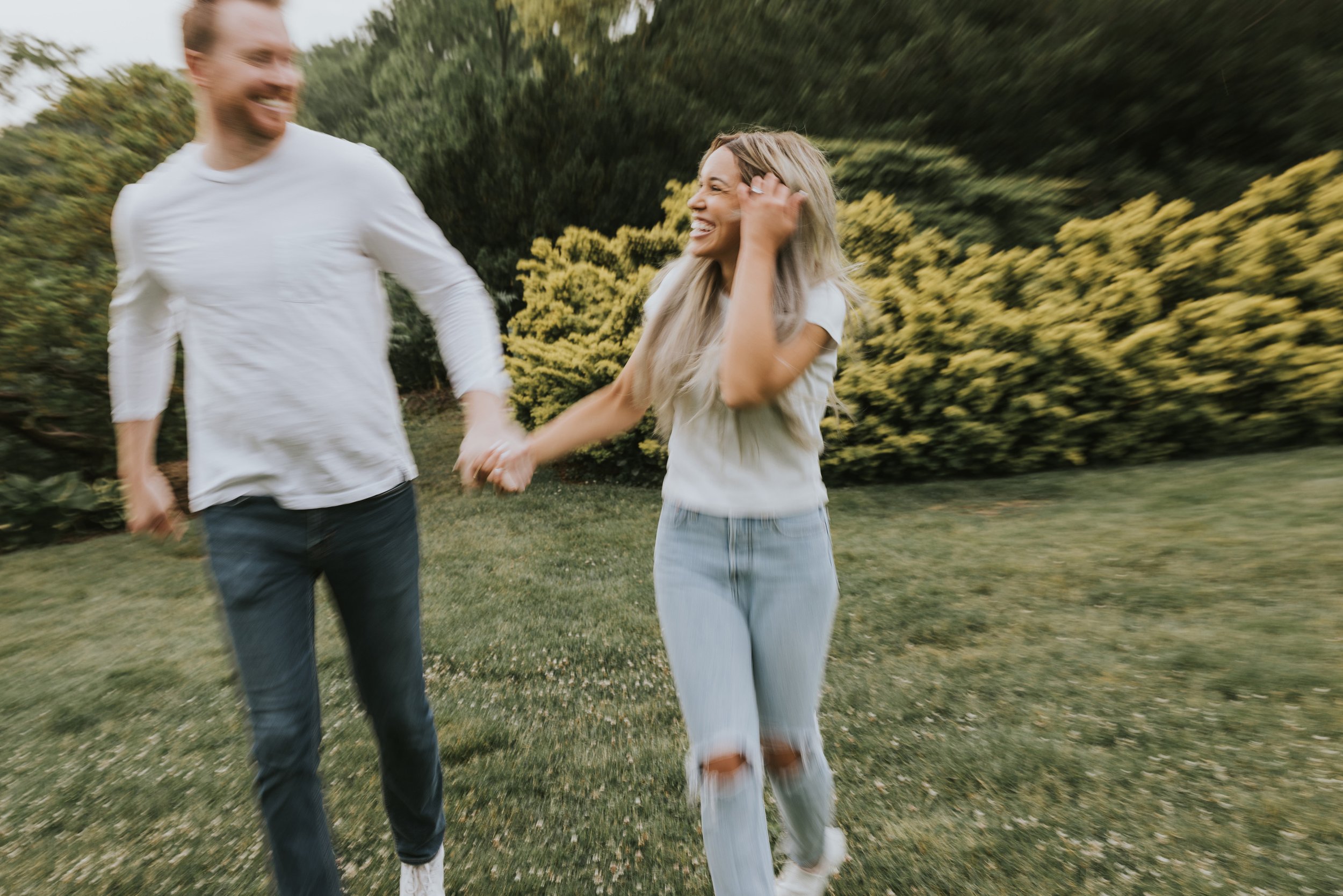 A playful couple runs and laughs together in a garden, capturing a joyful moment in motion during their engagement session.