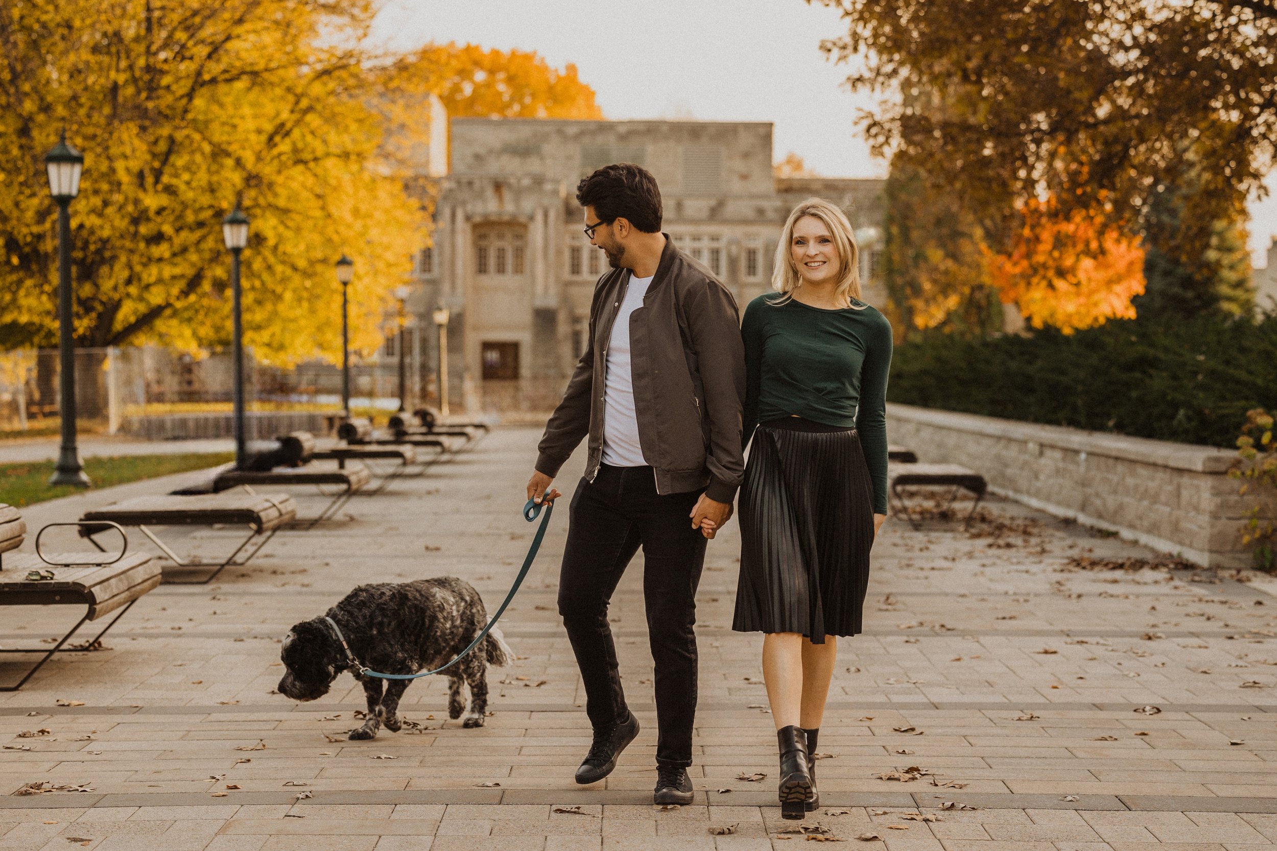 A couple walks hand in hand with their dog on a leaf-strewn path during an autumn engagement session. The vibrant fall colors create a picturesque and romantic atmosphere.