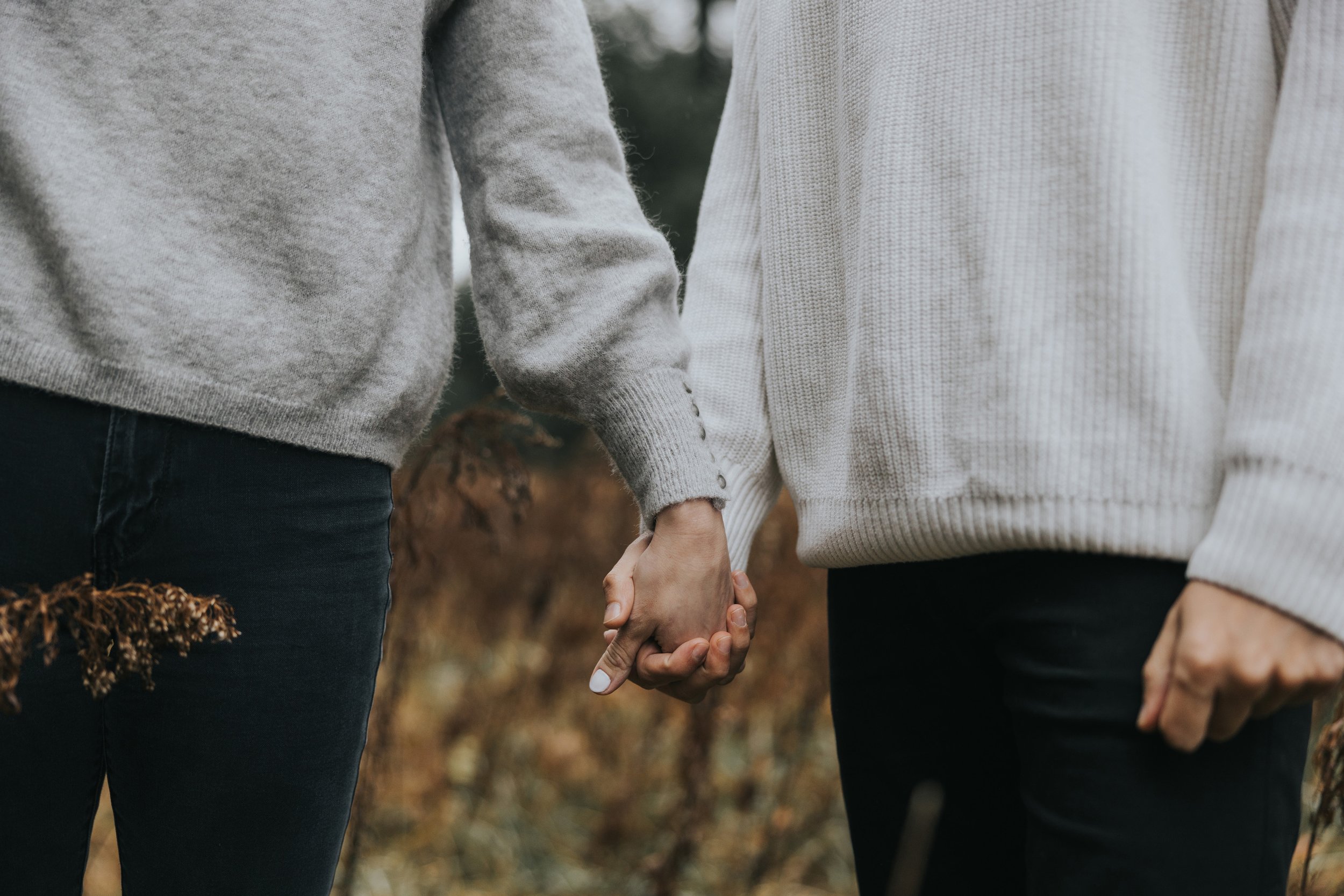 A close-up of a couple holding hands in an autumn meadow, both dressed in cozy sweaters, capturing a tender moment.