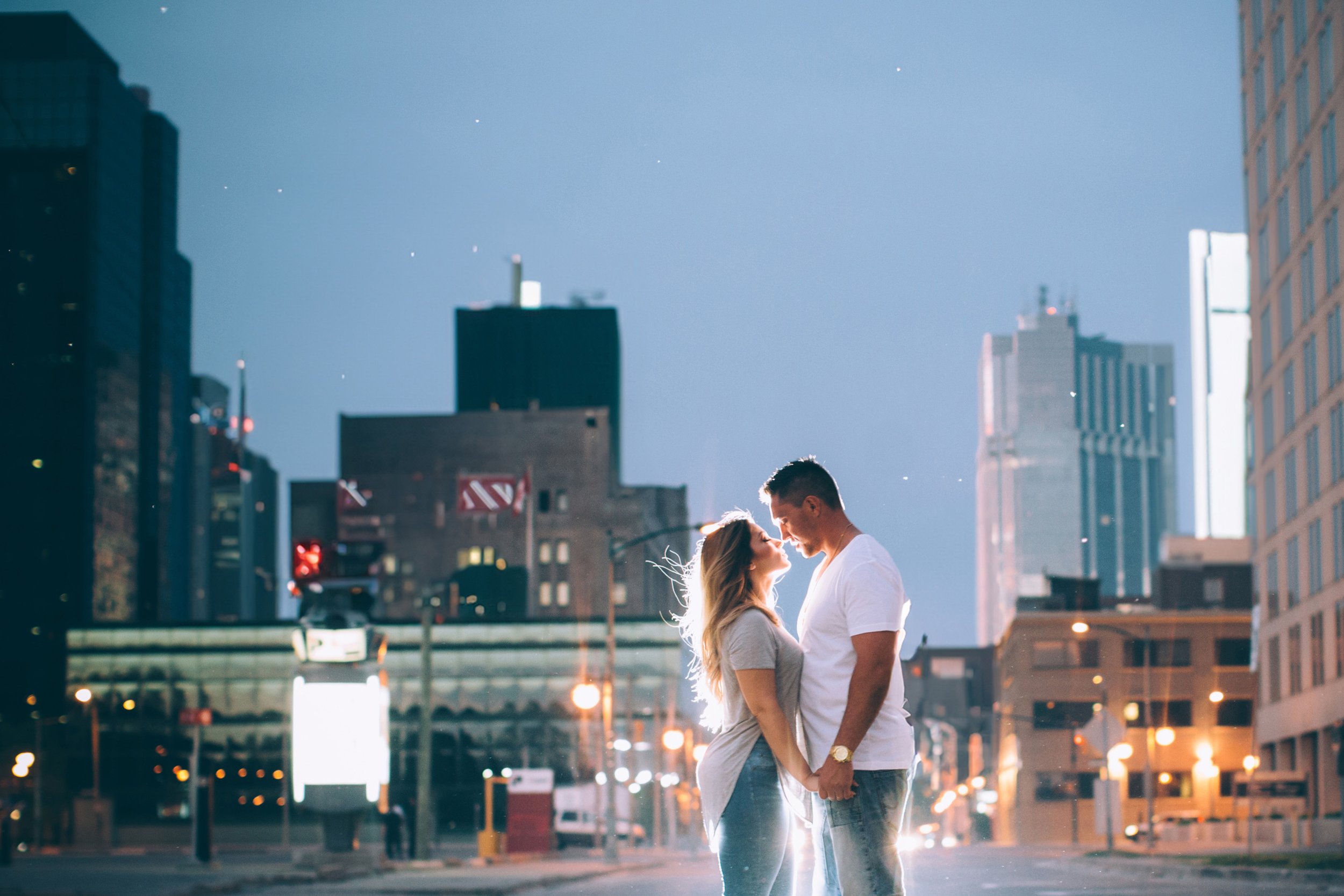 An evening engagement portrait of a couple in front of a city skyline, with soft light highlighting their embrace against the backdrop of urban lights.