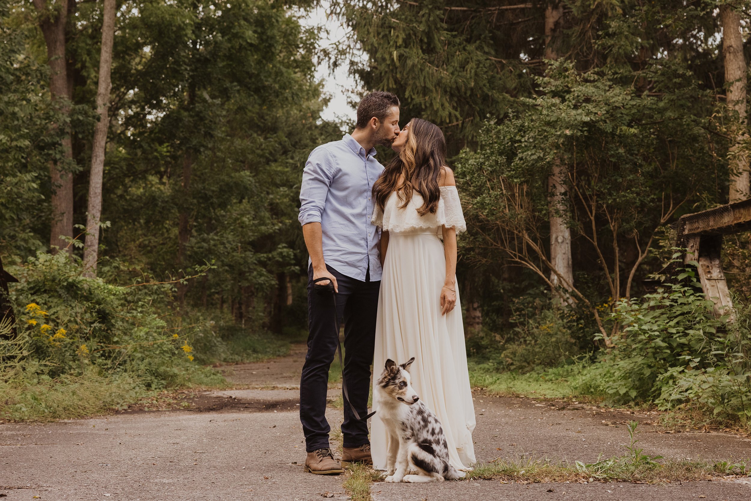 A couple shares a kiss while standing in a forest path, accompanied by their dog, during a romantic engagement session in London, Ontario.