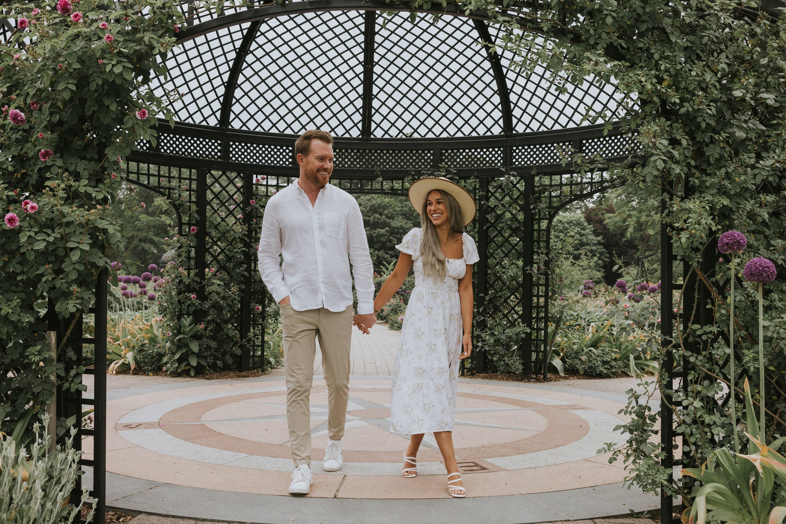 A couple holding hands and walking through a garden under an arbour adorned with flowers.