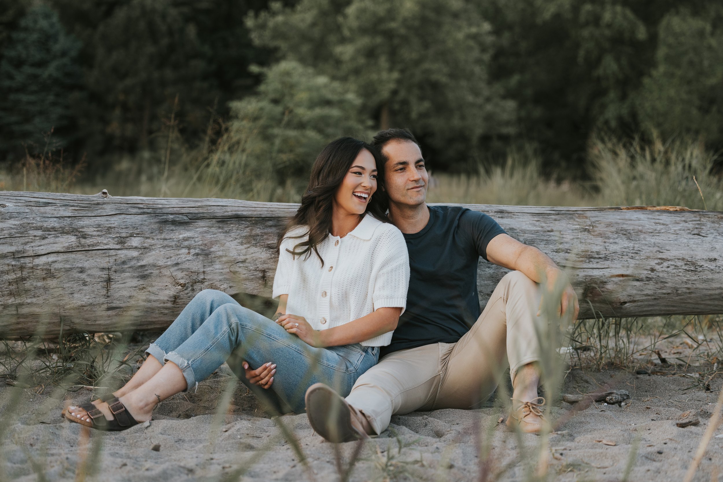 A couple relaxes and laughs together while sitting on a beach, leaning against a weathered wooden log, with nature surrounding them.