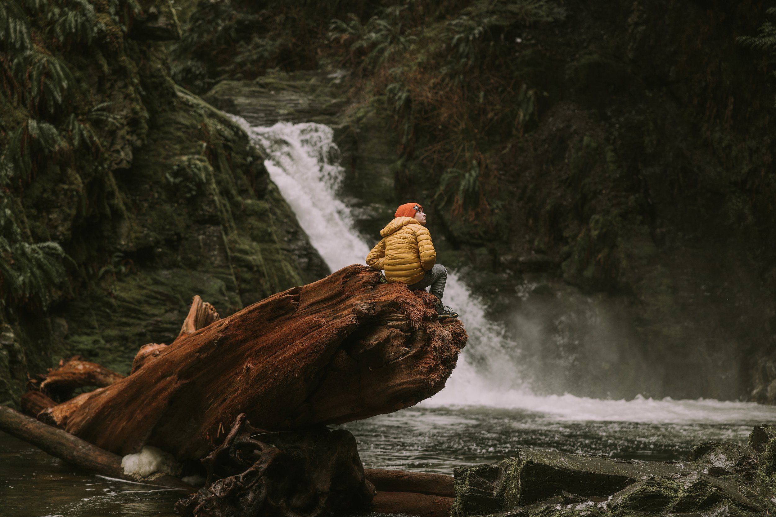 A boy wearing a bright yellow jacket sits on a large rock overlooking a flowing waterfall, embracing the serenity of nature during a hike.