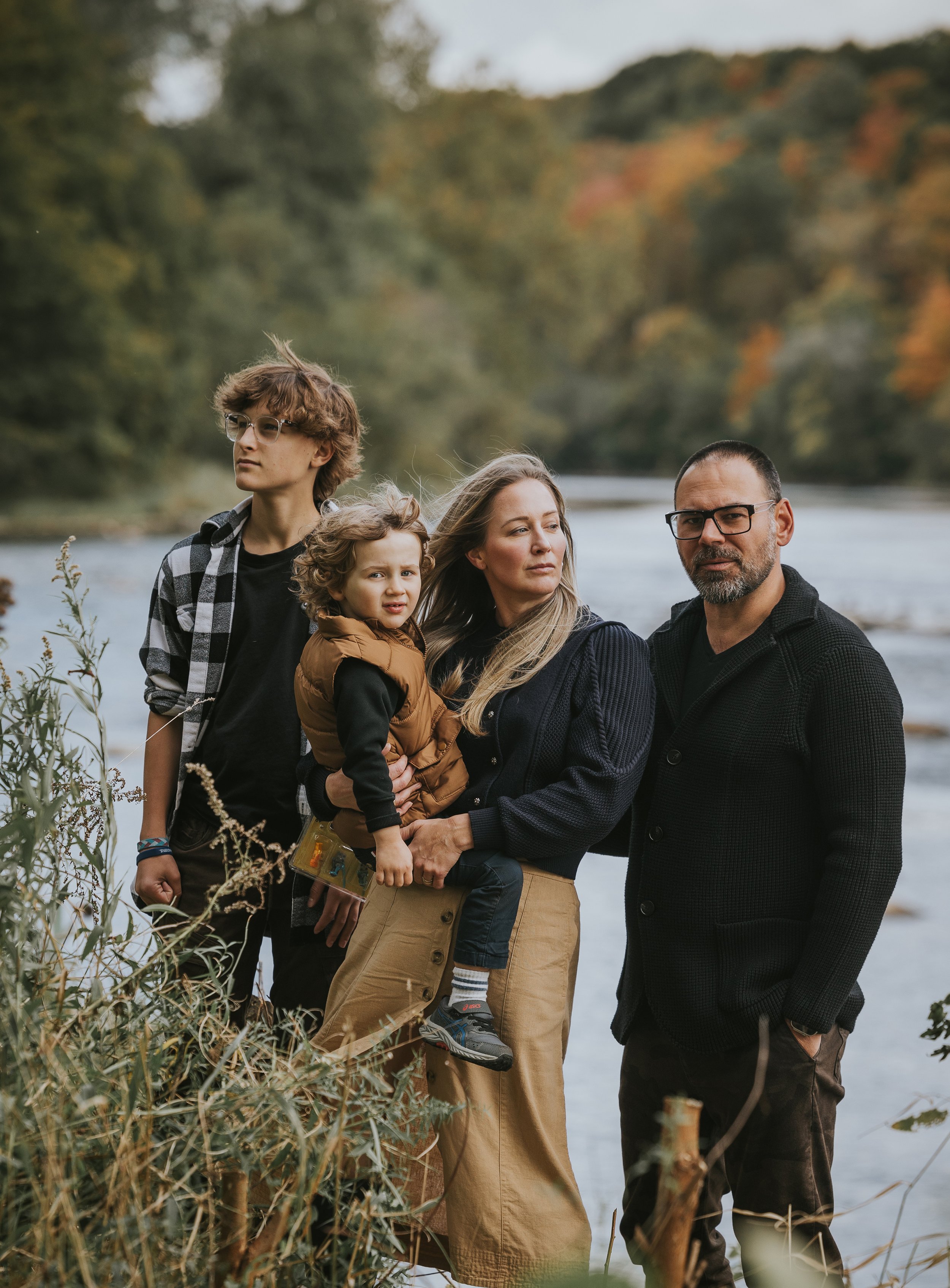 A family of four standing by a river with autumn trees in the background, dressed warmly and gazing thoughtfully into the distance.