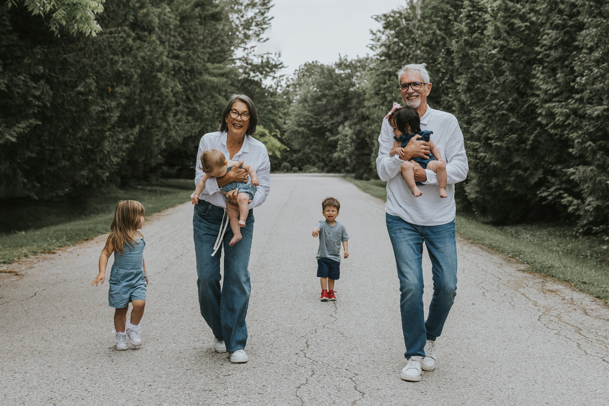 Grandparents joyfully walking down a tree-lined path with their young grandchildren, each carrying a baby.