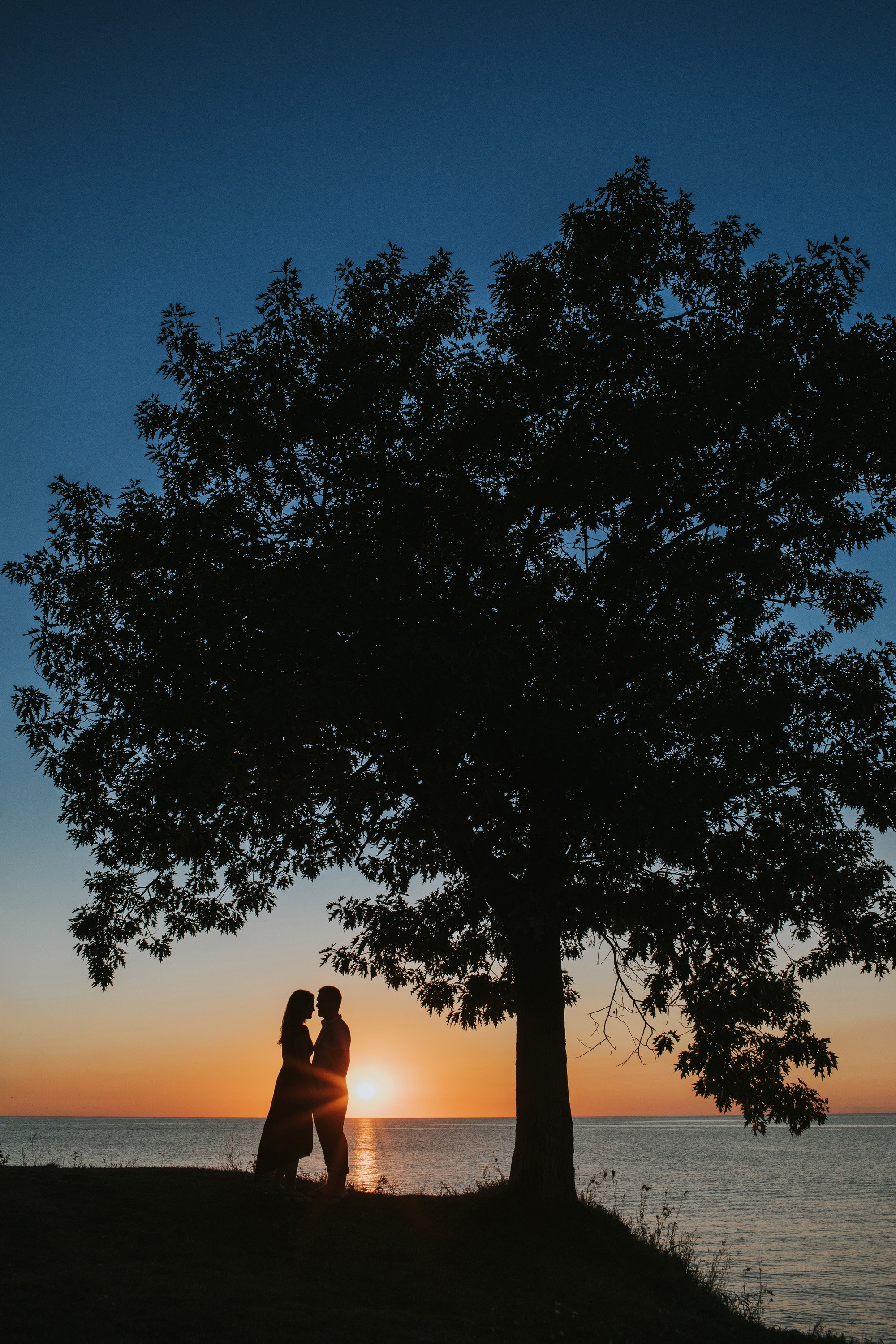 A couple standing beneath a tree, silhouetted against a vibrant sunset over a lake, sharing a quiet, intimate moment.