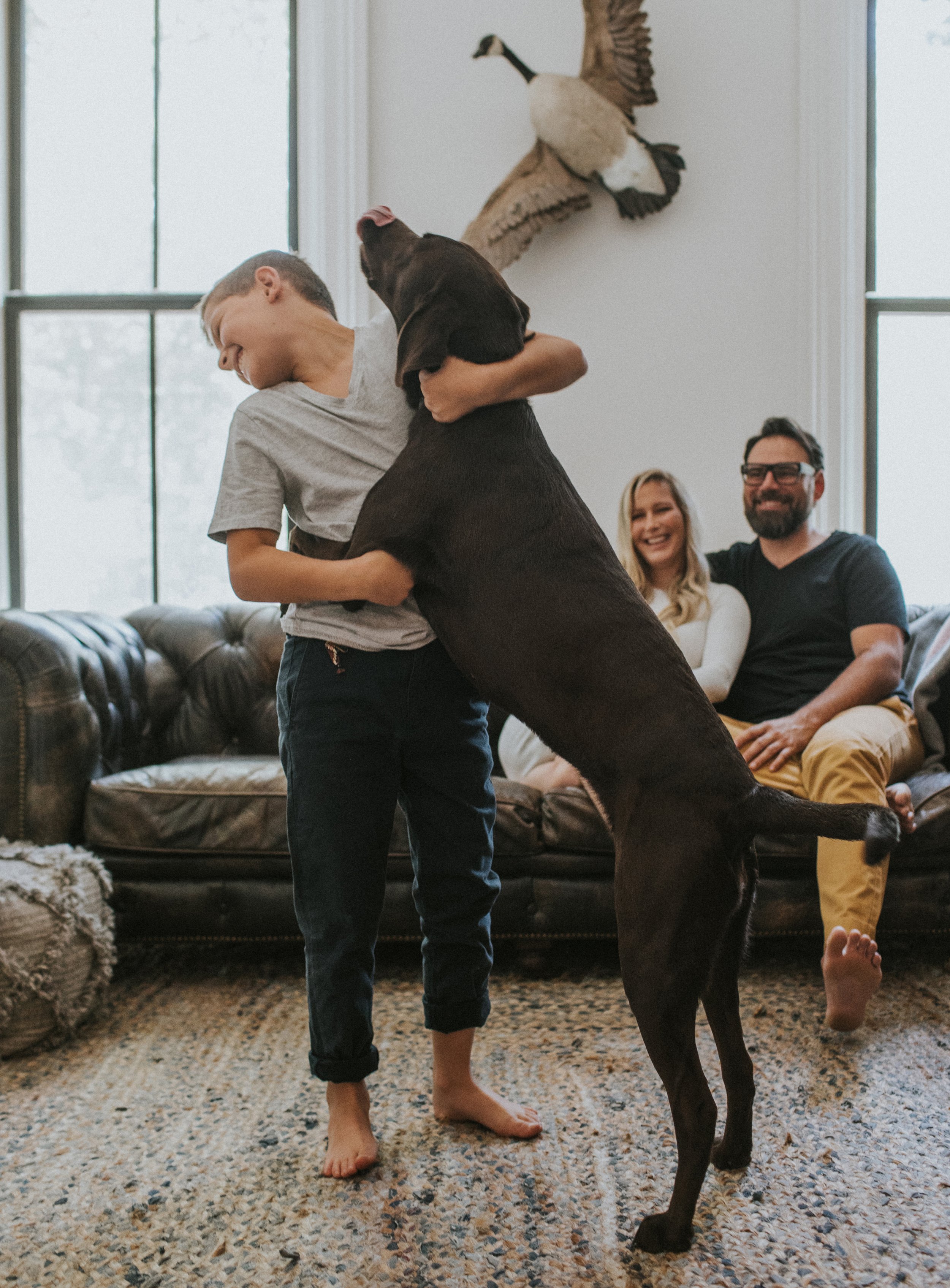 A young boy playfully hugging his dog in the living room while his parents smile in the background.