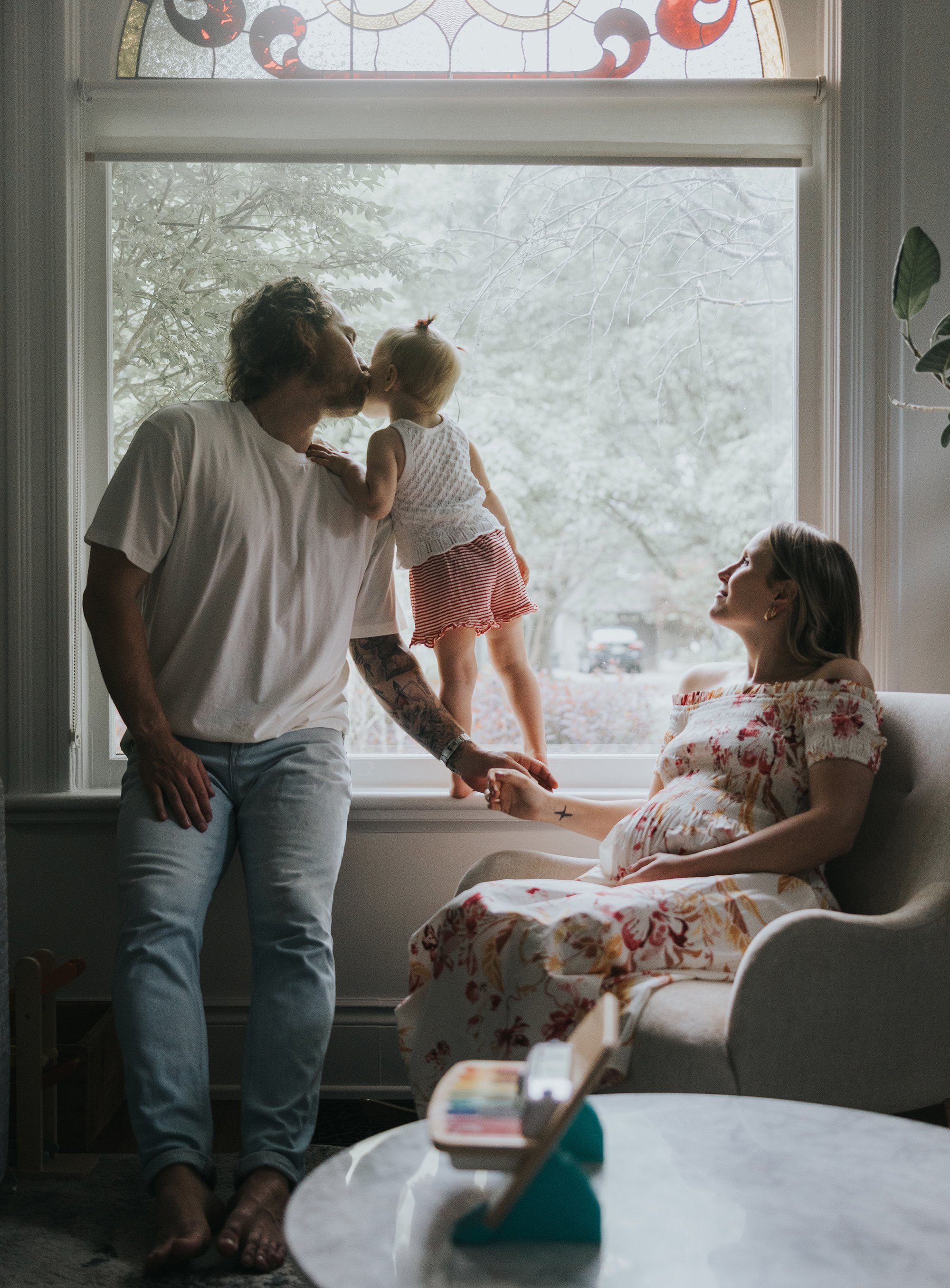 A sweet moment where a father kisses his young daughter on the head while the mother, expecting a new baby, looks on lovingly at home.