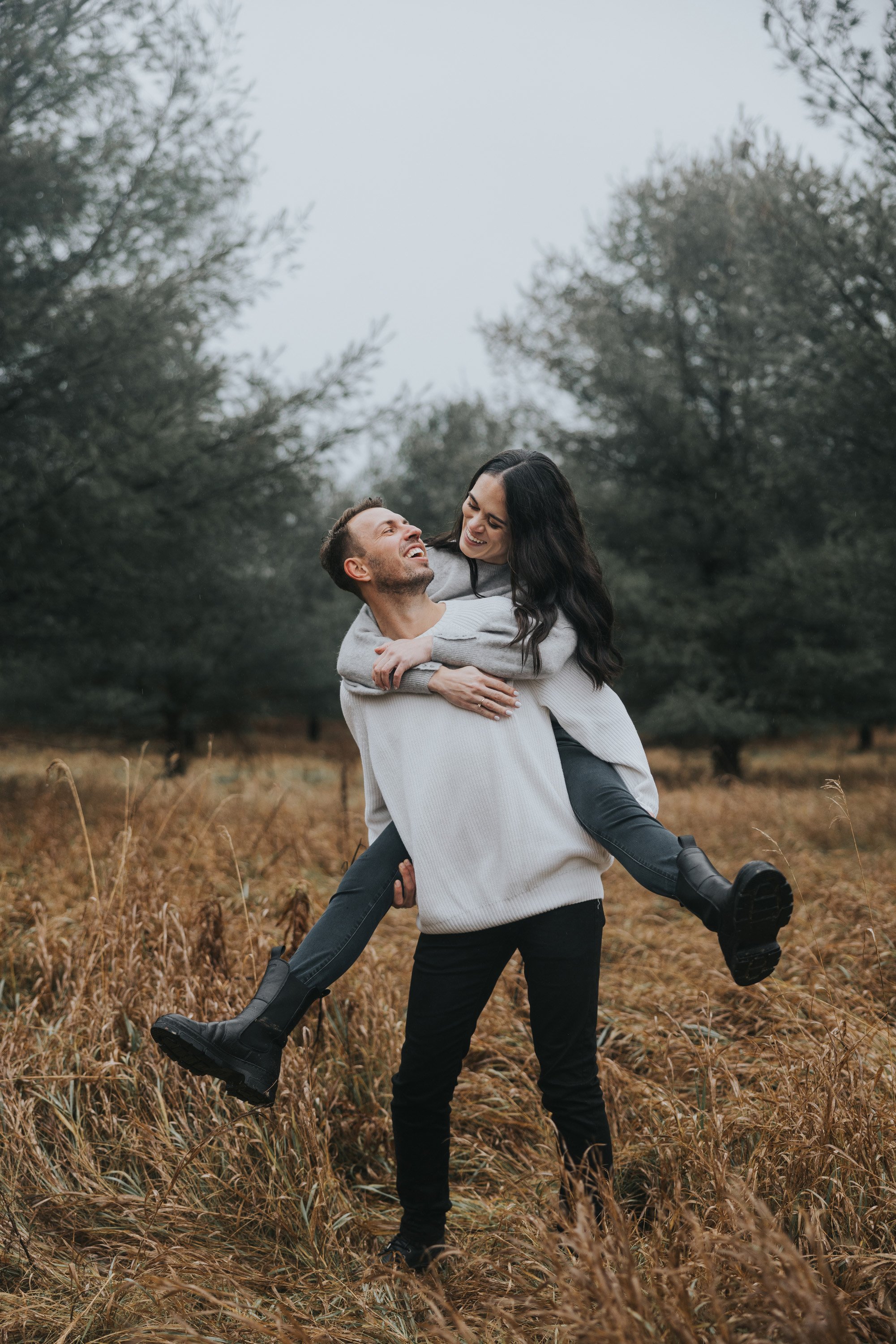 Playful engagement session with a couple laughing as the woman gives a piggyback ride in a forest setting.