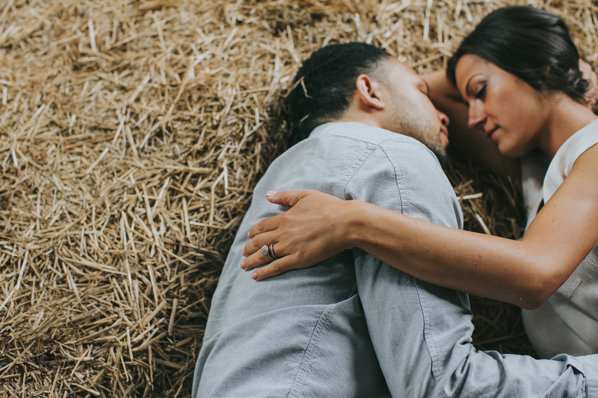 A couple lies close together on a bed of hay inside a rustic barn, sharing an intimate moment during their engagement session, with the focus on the woman’s hand showing off her engagement ring.